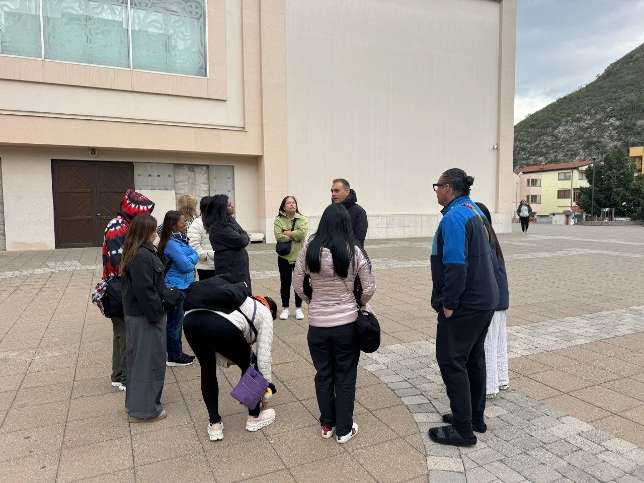 Group of people standing in a circle outside, listening to a man in a black jacket, in front of a large beige building with windows.