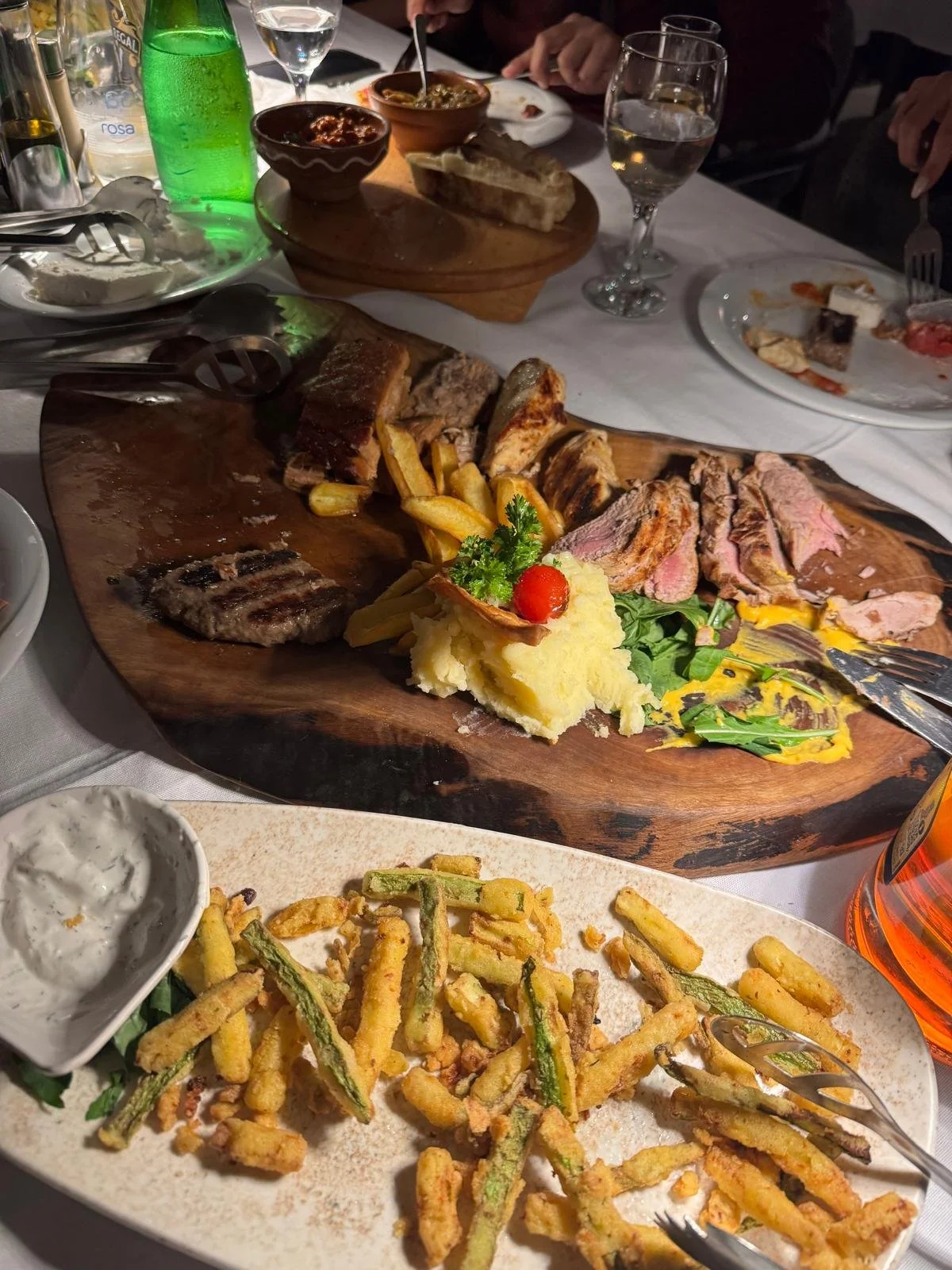 A table set with a variety of food including a large wooden platter of sliced beef and lamb, mashed potatoes with garnish, and a plate of fried vegetables with dipping sauce.