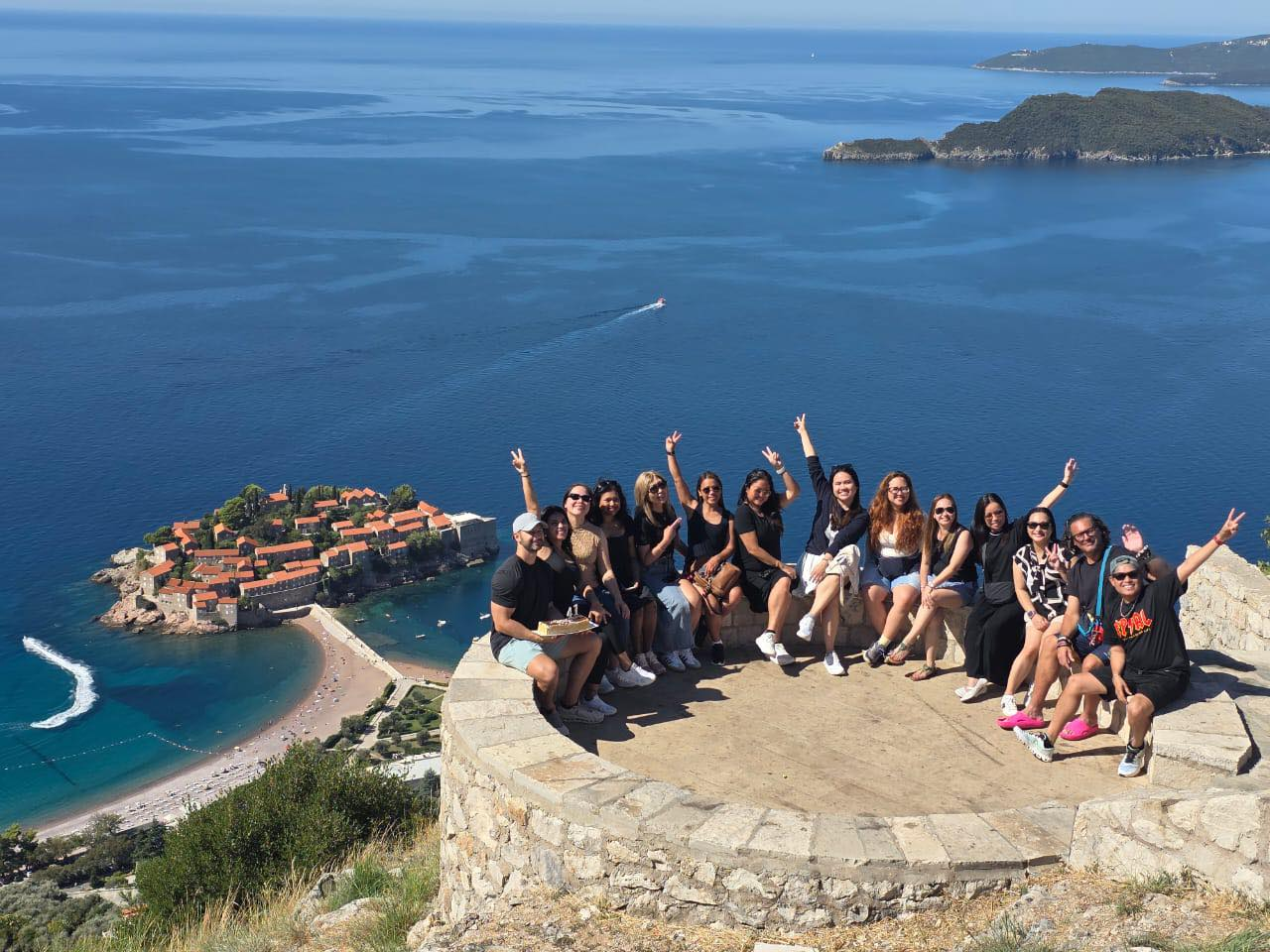 Group of thirteen people sitting on a stone platform with the ocean and island in the background, making peace signs and smiling.