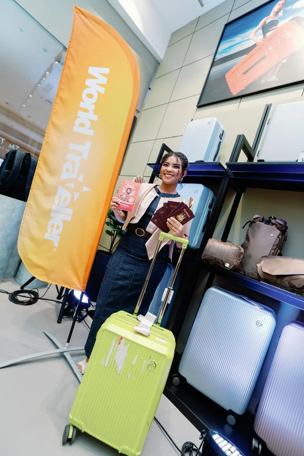 A smiling woman holding passports and an travel brochure standing with her yellow suitcase in front of an 'Amaland Travel' flag at an airport or travel store.