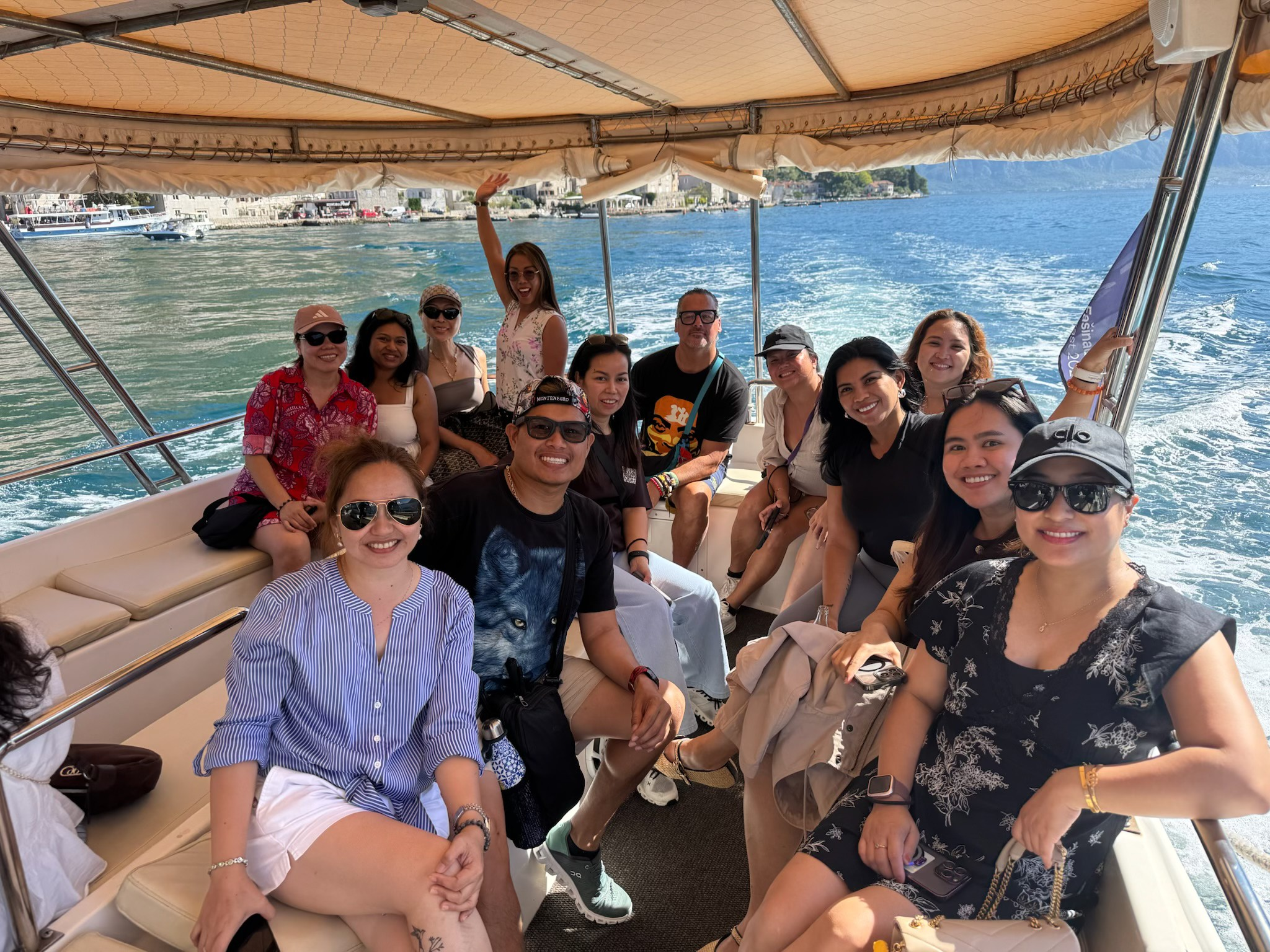 A group of people on a boat ride enjoying sunny weather, smiling for the photo, with water and a shoreline in the background.