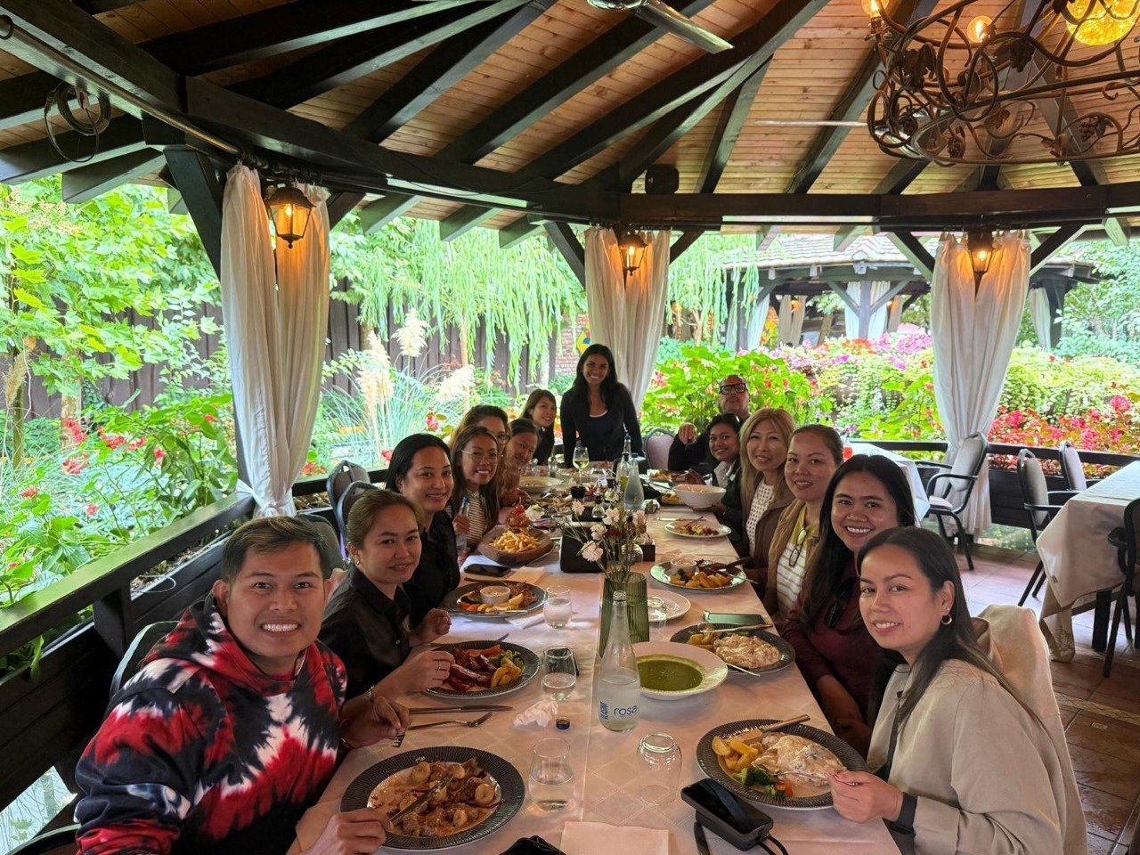 A group of people dining together at a long table in an outdoor restaurant with lush green plants and flowers in the background. The restaurant is covered with a wooden roof and has white curtains and elegant lighting.