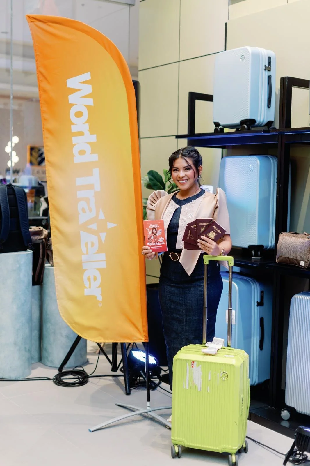 A woman at a travel exhibition holding brochures and a digital travel guide, standing with a green suitcase and a yellow feather flag that reads 'World Traveler', surrounded by luggage displays.