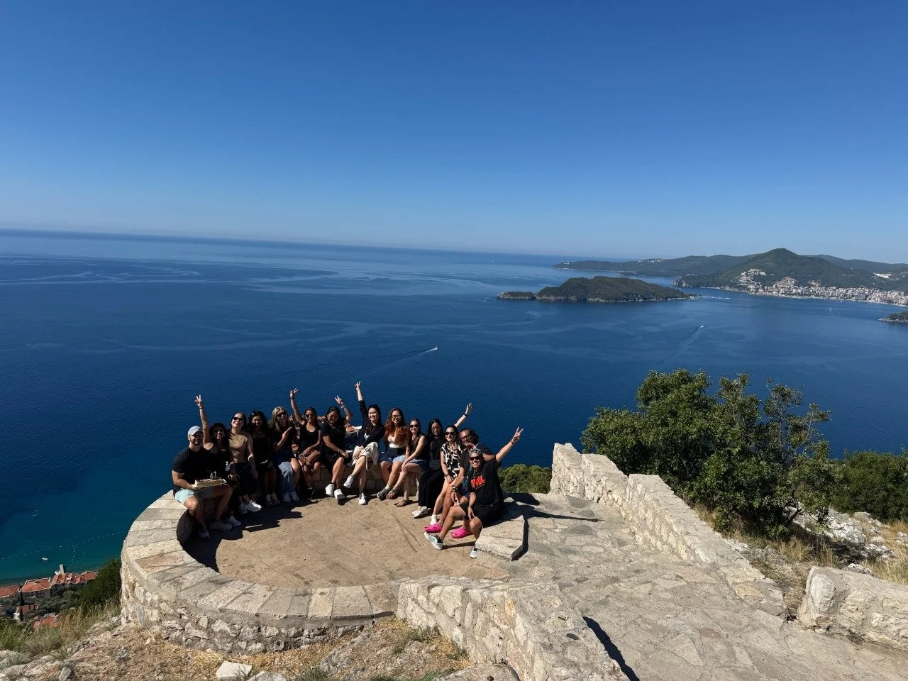 Group of people sitting and standing on a stone platform overlooking a large body of water with islands and hilly land in the background on a sunny day.