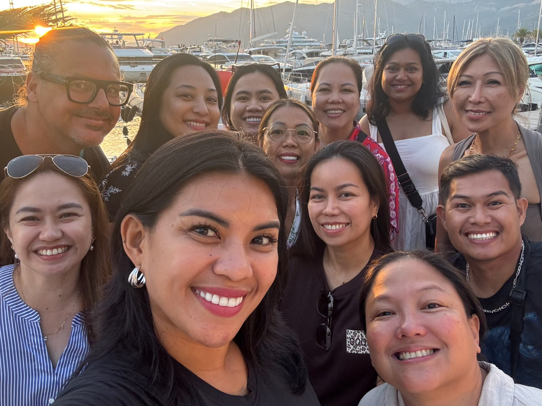 A group of people smiling for a photo at a marina during sunset, with boats docked in the background.