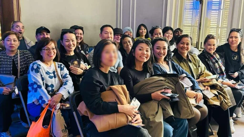 Group of women and men sitting in a room, smiling and looking towards the camera, during an indoor event or gathering.
