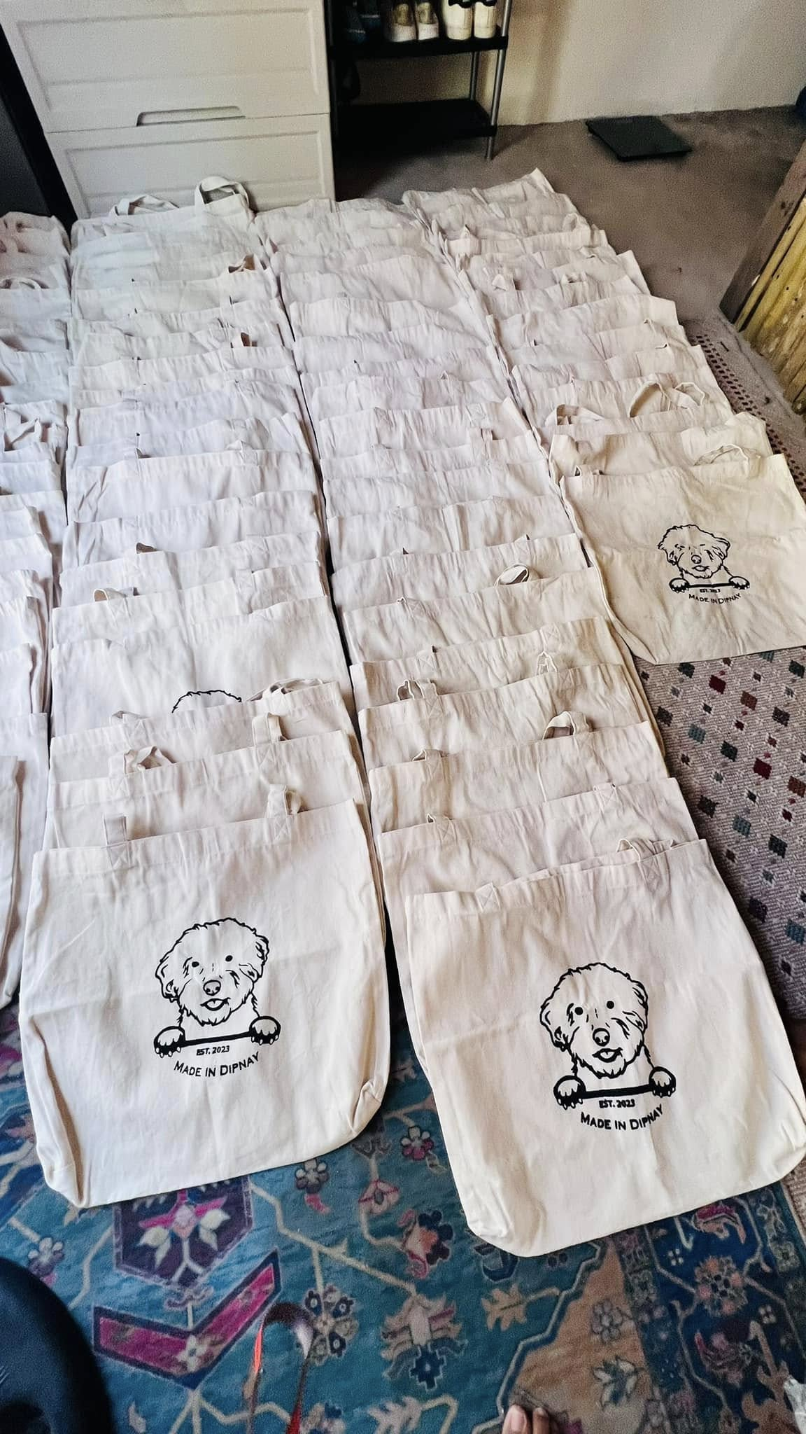 Numerous white tote bags with a black line drawing of a dog and text "EST. 2023 MADE IN DIPAY" printed on them, spread out across a carpeted floor.