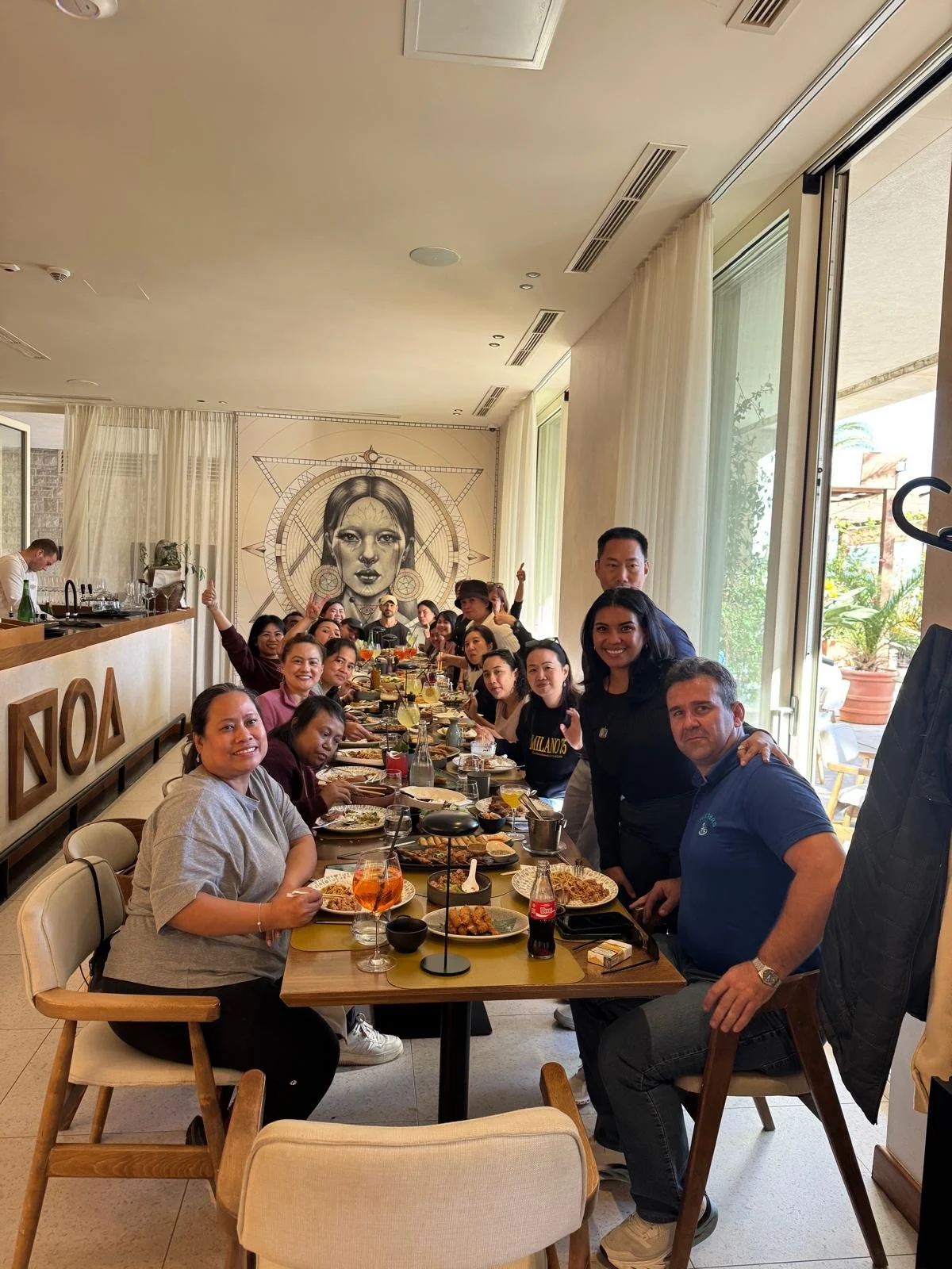 A group of people gathered around a long table in a restaurant, enjoying a meal. The setting is bright with large windows and artwork on the wall depicting a woman's face with concentric circular designs.