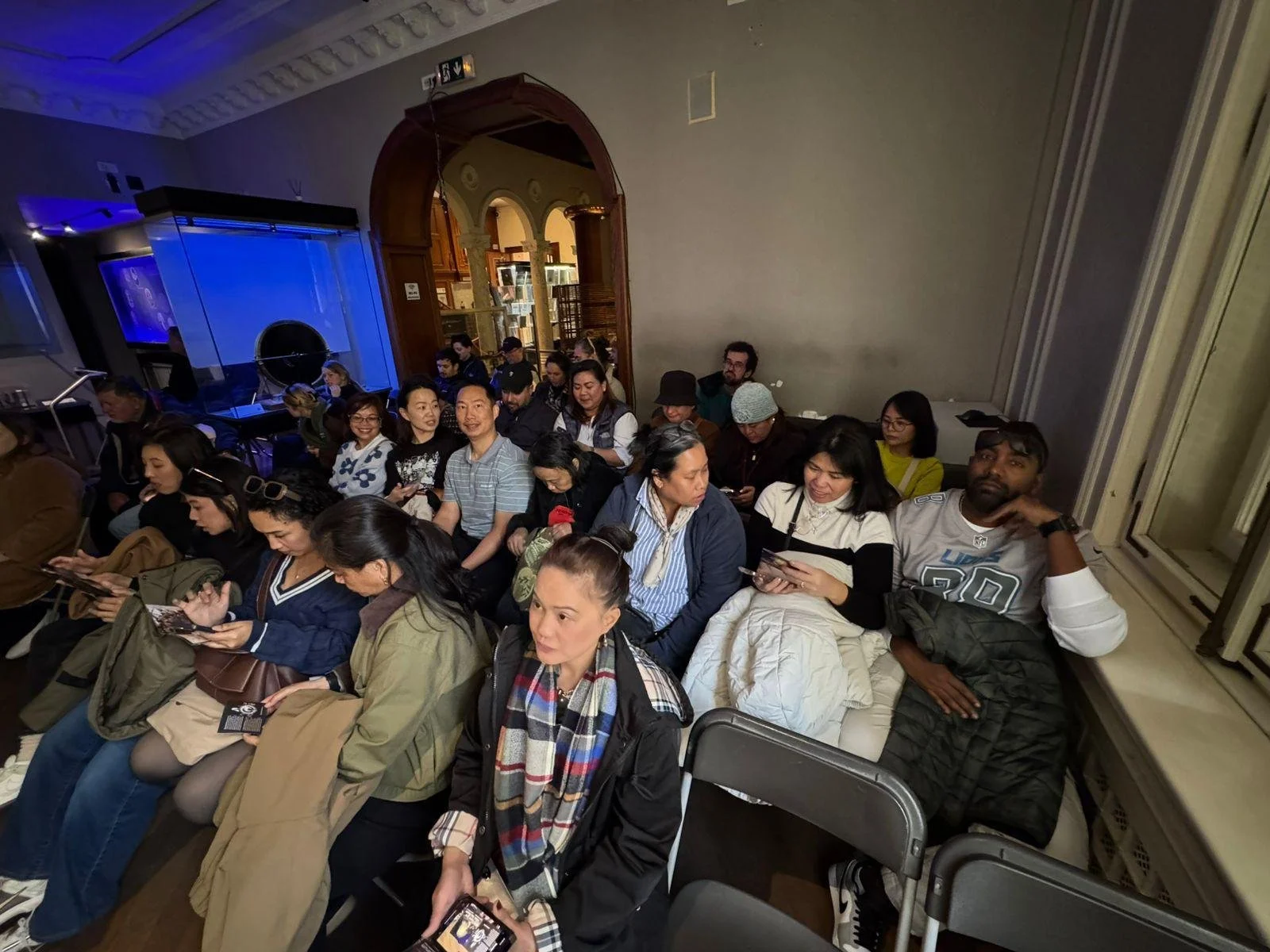 A diverse group of people seated indoors, many looking at their phones, with some engaging in conversation. There are blue lighting effects and a wooden archway in the background.