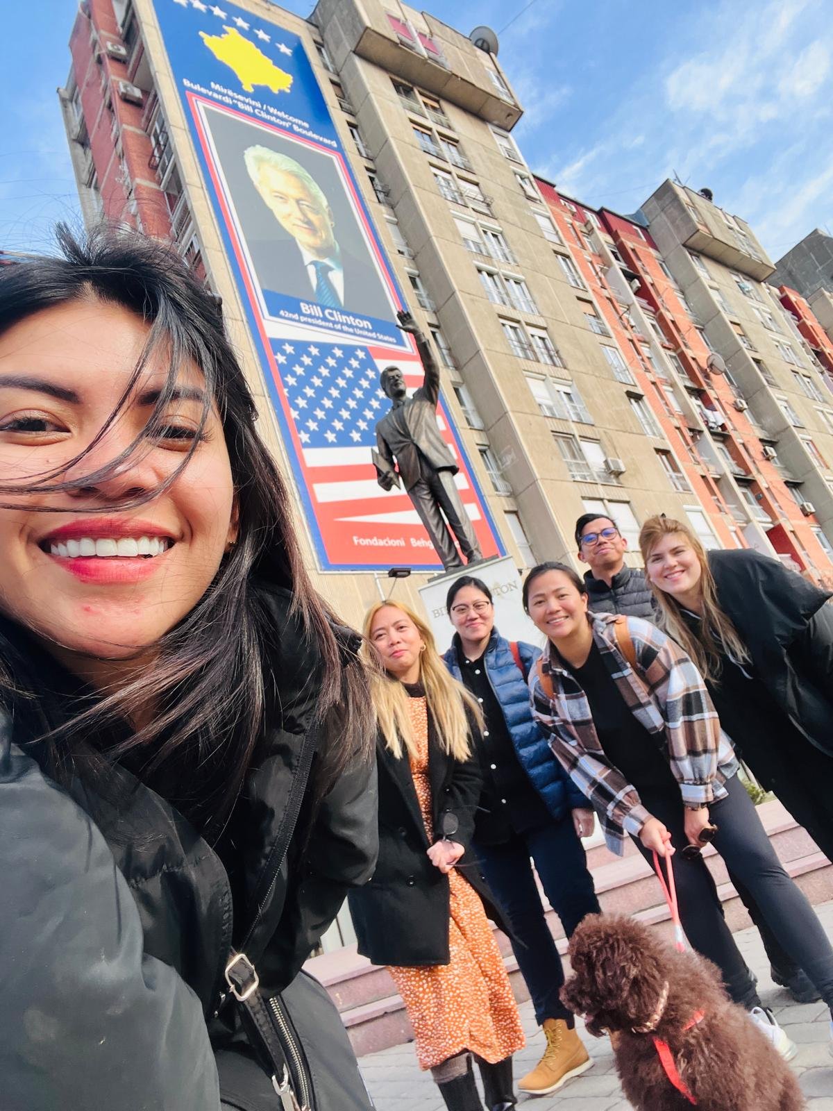 A group of six people and a dog taking a selfie outdoors in front of a large billboard featuring Bill Clinton and an American flag backdrop, with a statue and building in the background.