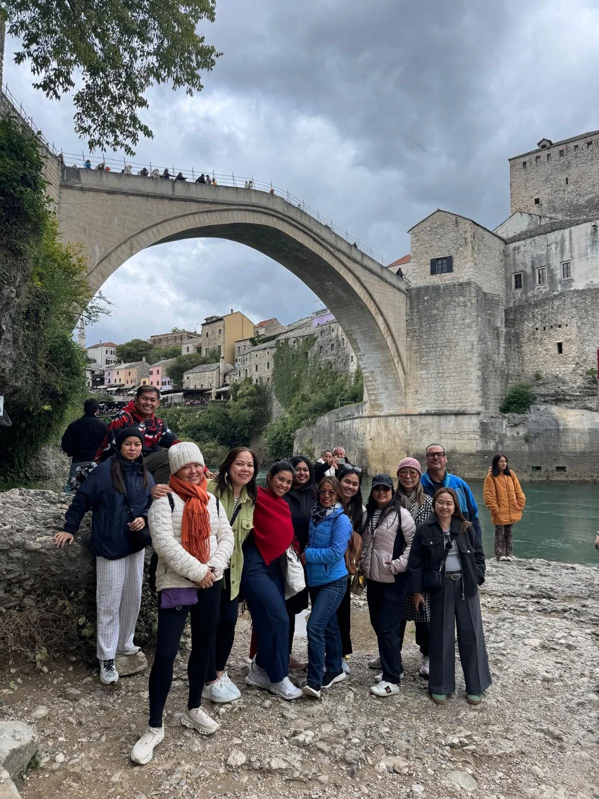Group of people posing for a photo on a rocky riverbank with the historic Ponte Vecchio bridge and old stone buildings in the background under cloudy skies.