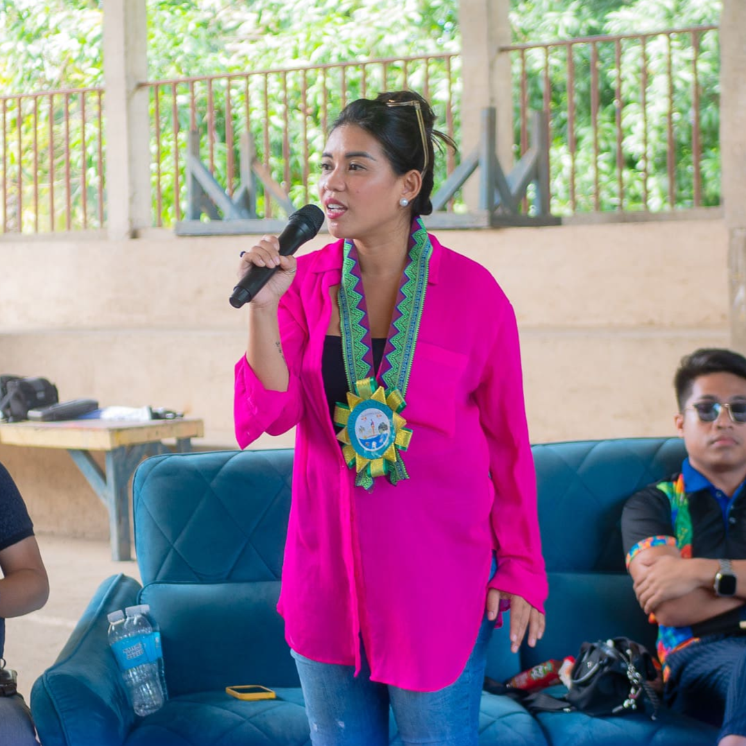 A woman wearing a bright pink shirt and a colorful necklace is speaking into a black microphone. She has sunglasses on her head and is standing beside a blue couch. There is a man sitting on the couch wearing sunglasses and a black shirt with colorful patterns, with his arms crossed. A small table with bottled water, a camera, and a smartphone is visible in the background, with greenery outside.