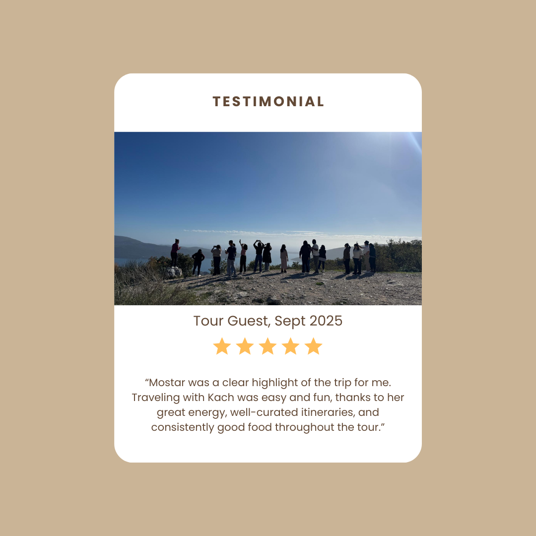 A group of people standing outdoors on a dirt path with a scenic view of mountains and a body of water under a bright blue sky, some taking pictures, during daytime.