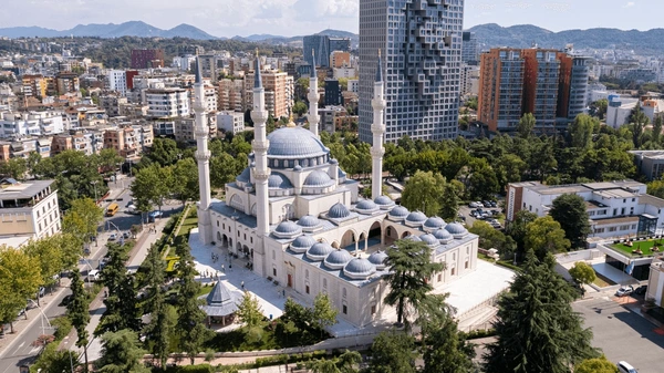 A white mosque with multiple domes and minarets surrounded by trees in an urban cityscape with high-rise buildings.