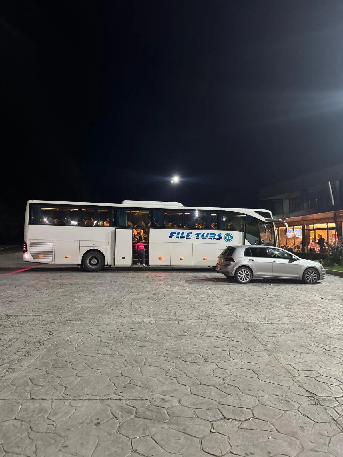 A white tour bus with blue lettering parked next to a silver car at night. Interior lights illuminate the bus, and people are visible inside. The surroundings include a building with some people outside.