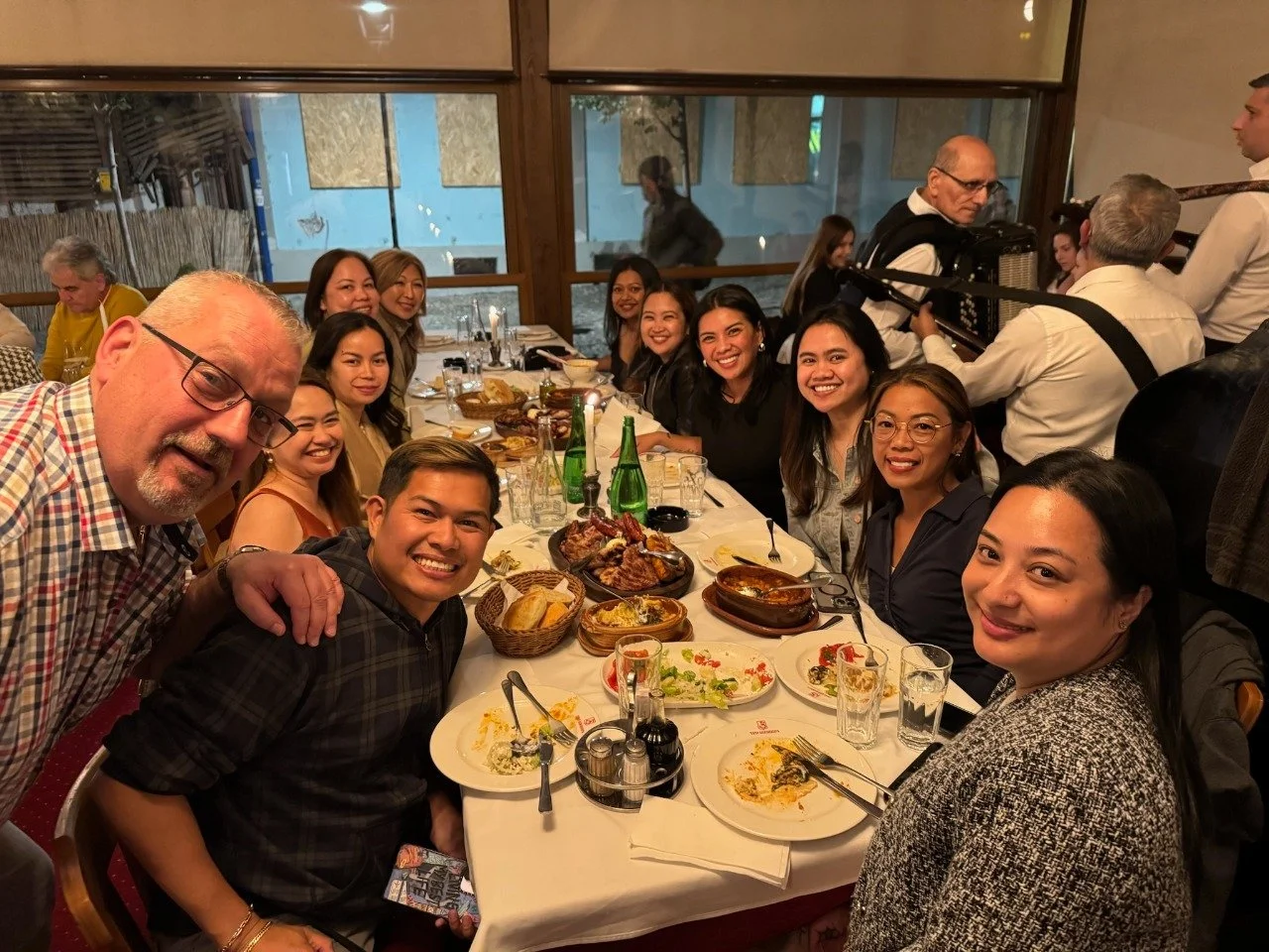 Group of people sitting at a long dinner table with food, smiling and posing for a photo. The table has plates, drinks, and cooked dishes, with a window and restaurant interior in the background.