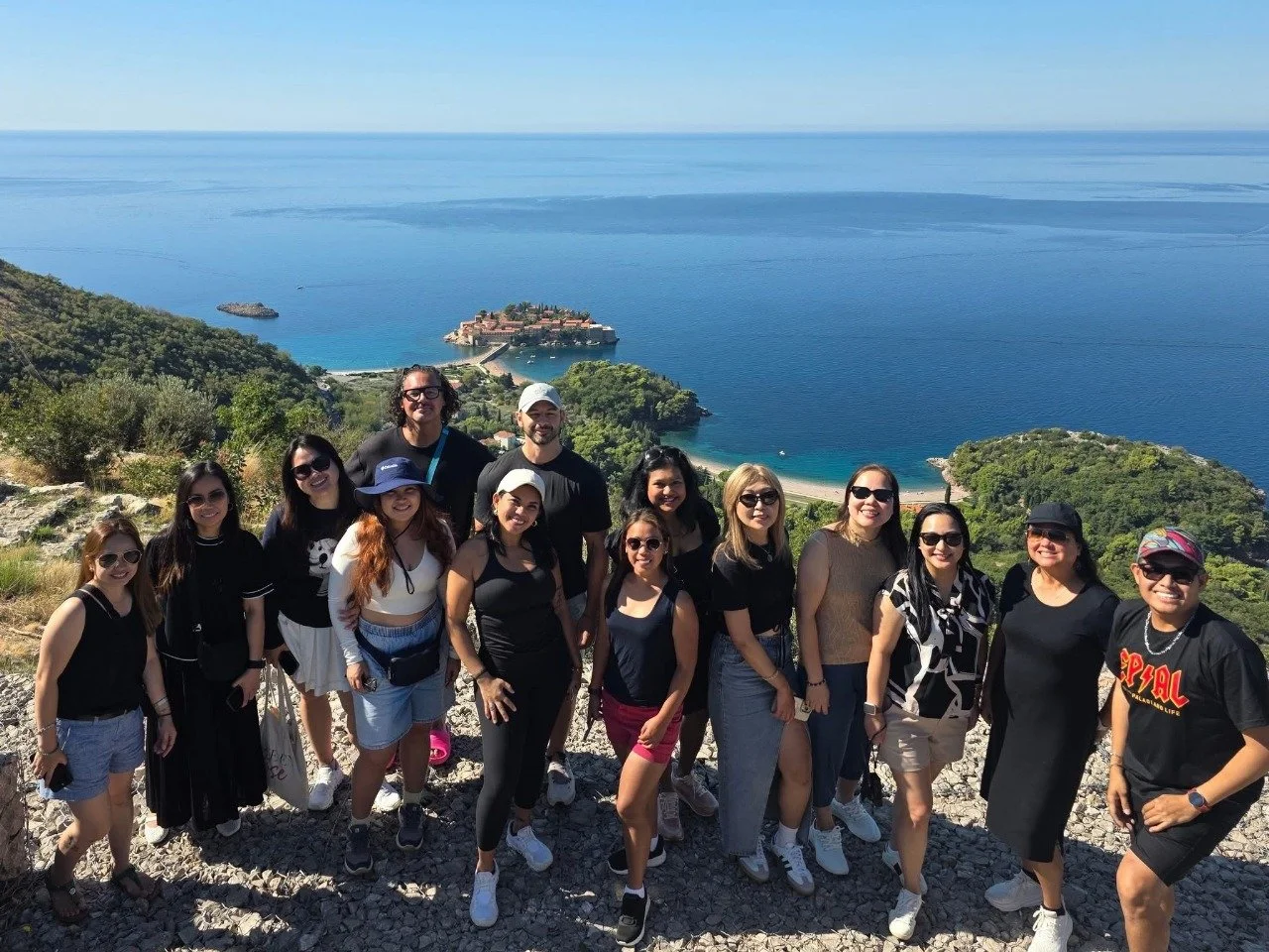 Group of diverse people posing together outdoors with a scenic ocean view, green hills, and a small island in the background in Budva, Montenegro.