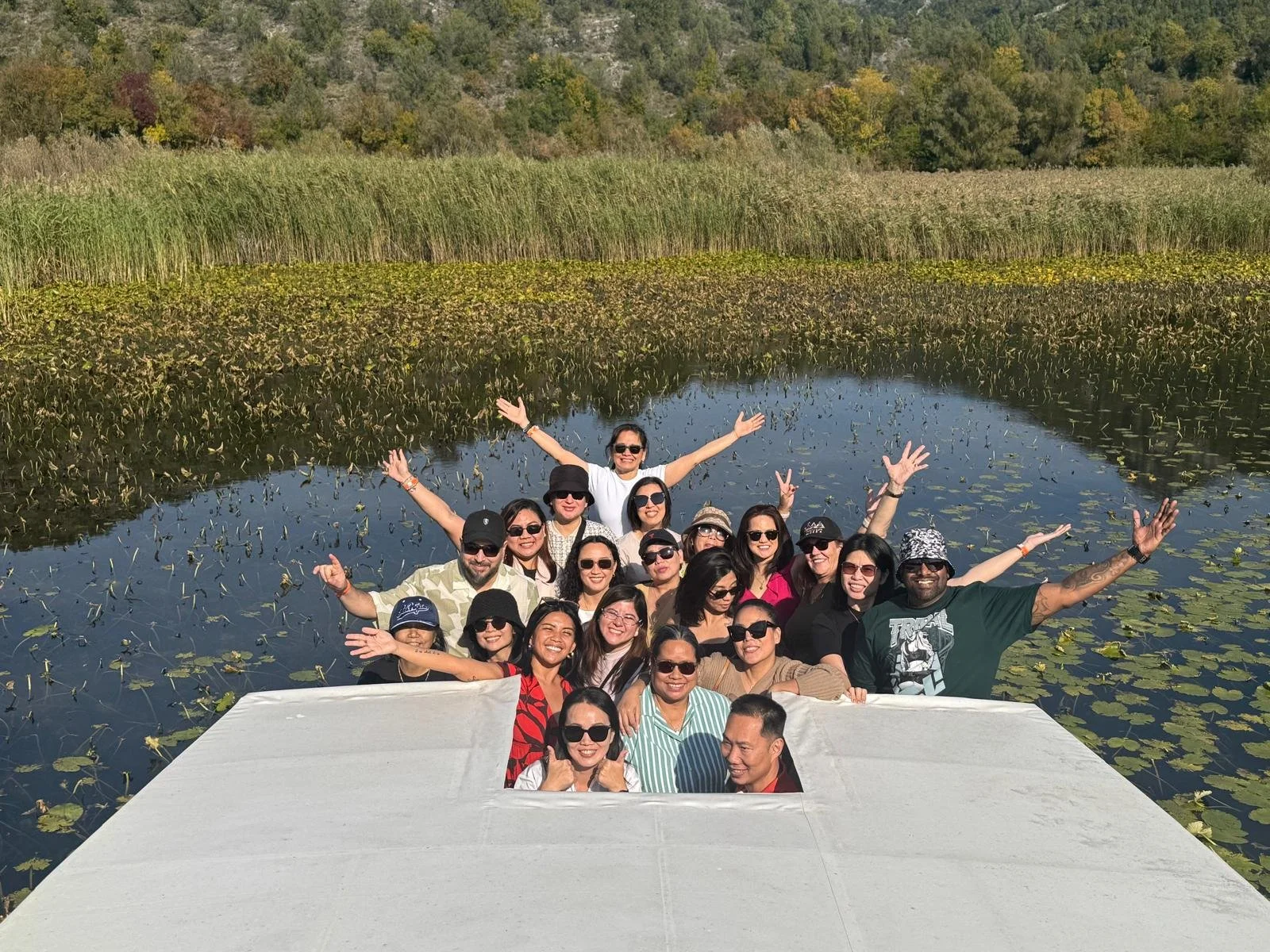 Group of smiling people on a boat in a pond with lily pads, surrounded by tall grass and trees, enjoying sunny weather.