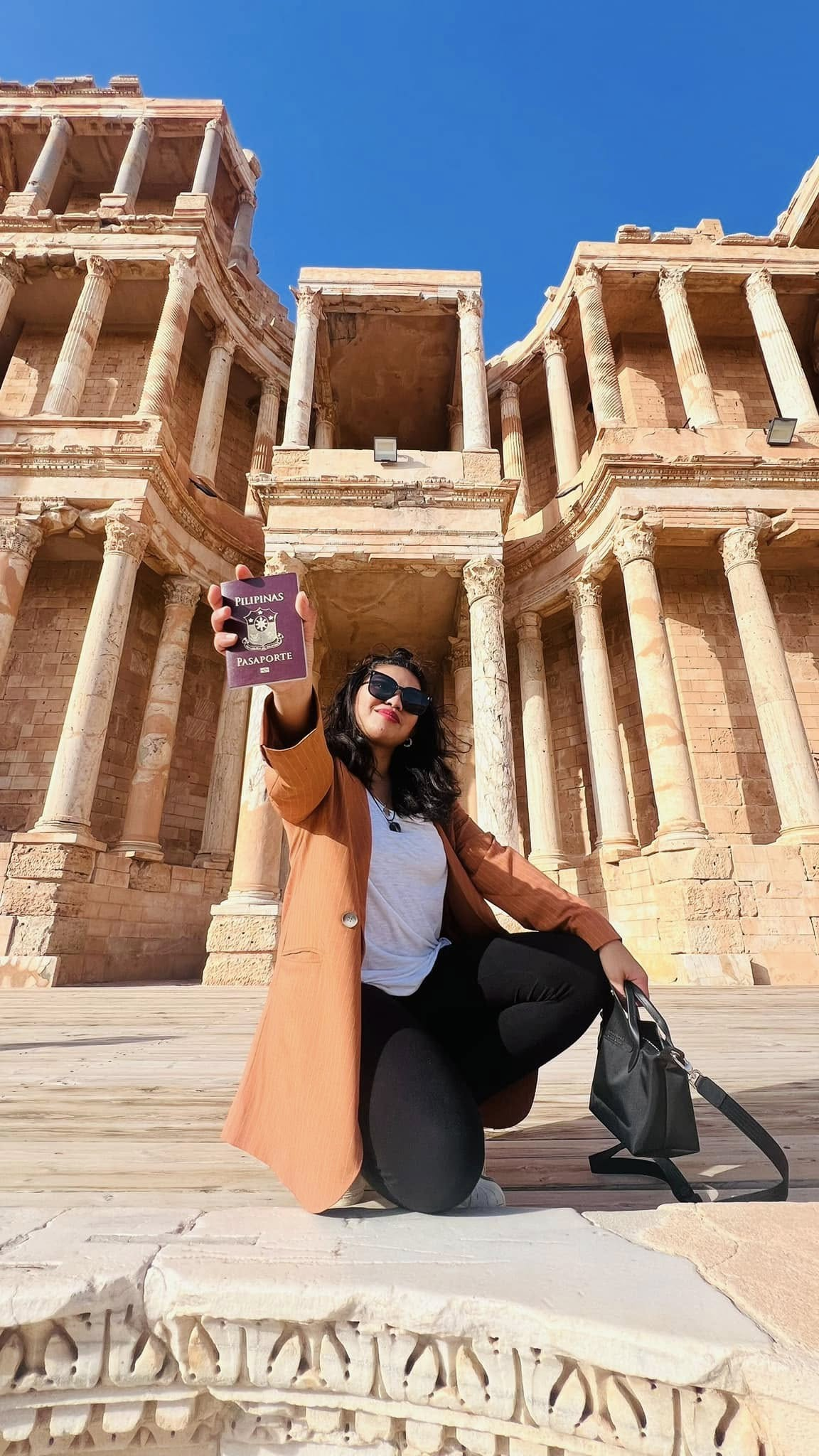 A woman with dark hair, sunglasses, and a brown coat holding a Philippine passport, squatting in front of an ancient Roman-style amphitheater or coliseum with columns under a clear blue sky.