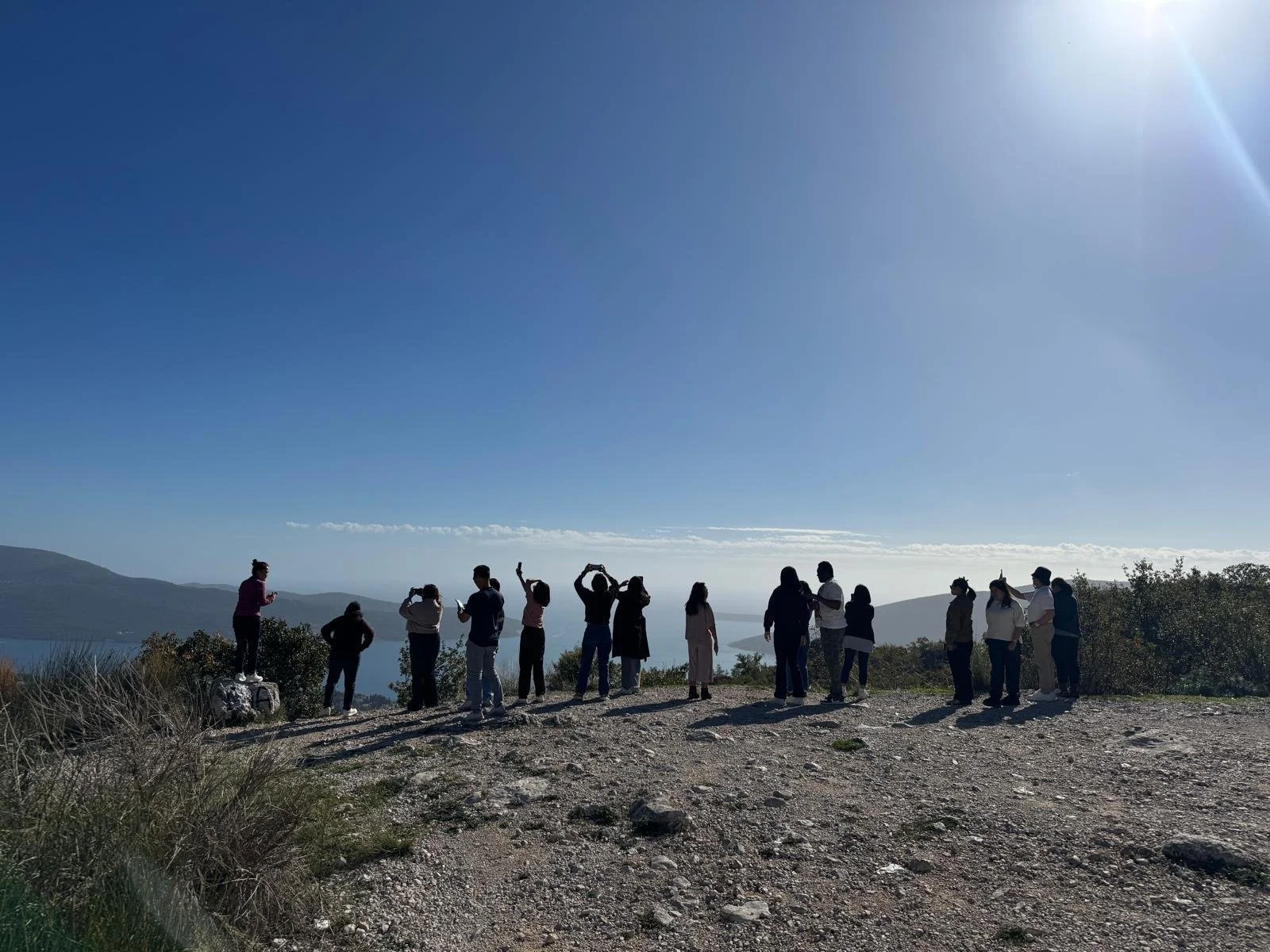 A group of people standing on a rocky hilltop in the border town of Herceg Novi, Montenegro overlooking a scenic landscape with distant hills, water, and a clear blue sky with bright sunlight.