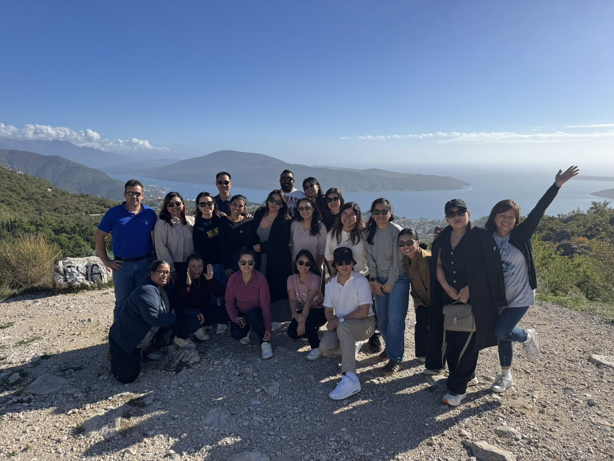 Group of people posing outdoors on a mountain with a lake and hills in the background, some smiling and others raising their hands.