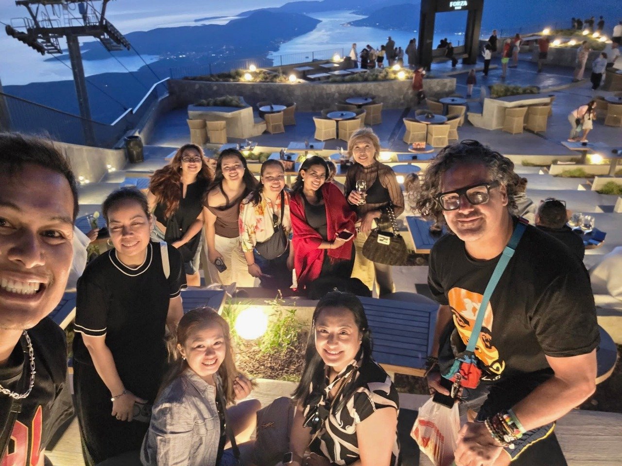 Group of people smiling at camera during an evening gathering on a rooftop terrace with a view of a mountain and water in the background on top of the cable car in Kotor, Montenegro