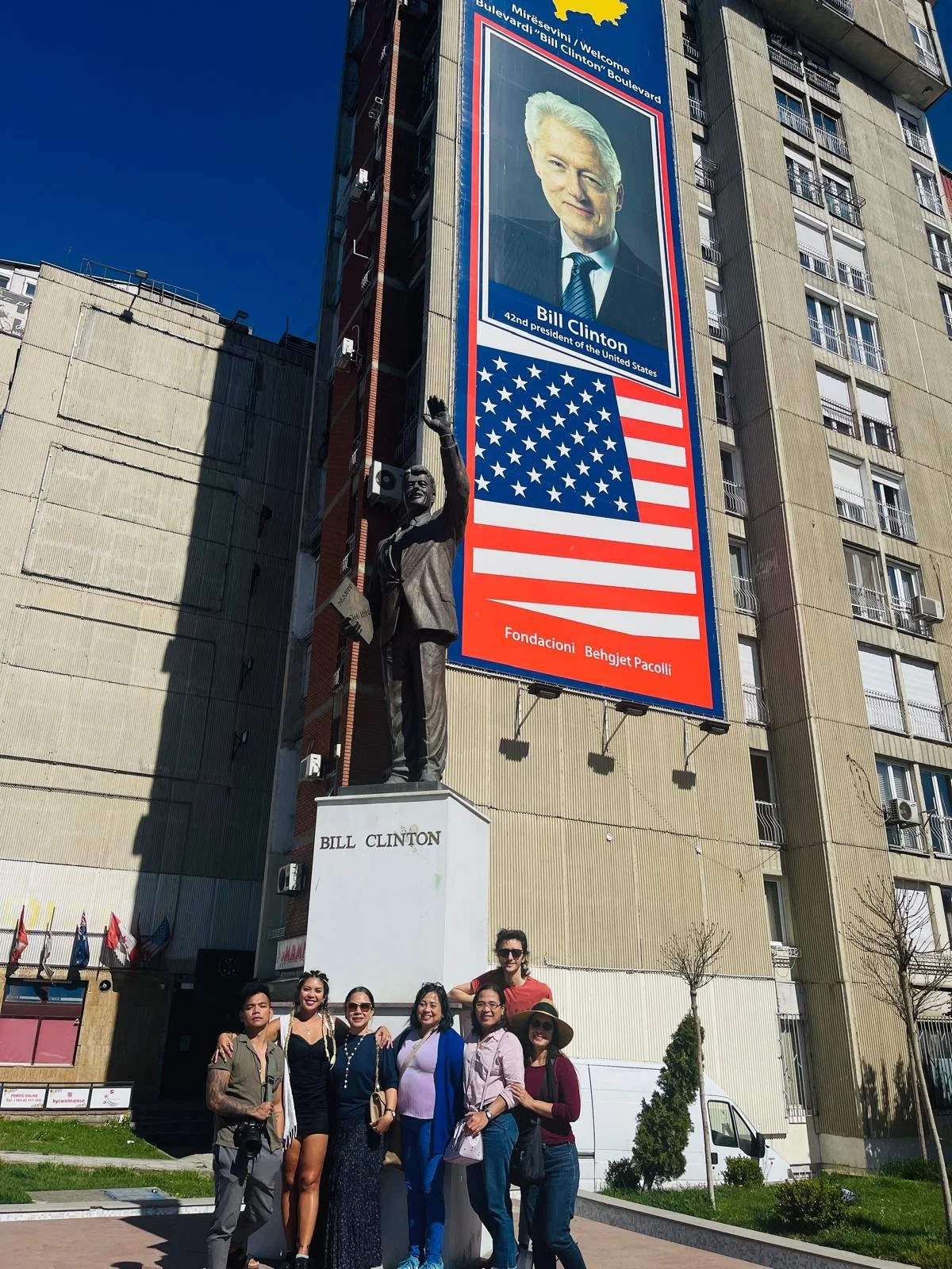 Group of six people standing in front of a statue of Bill Clinton, with a large billboard of Bill Clinton and an American flag in the background.