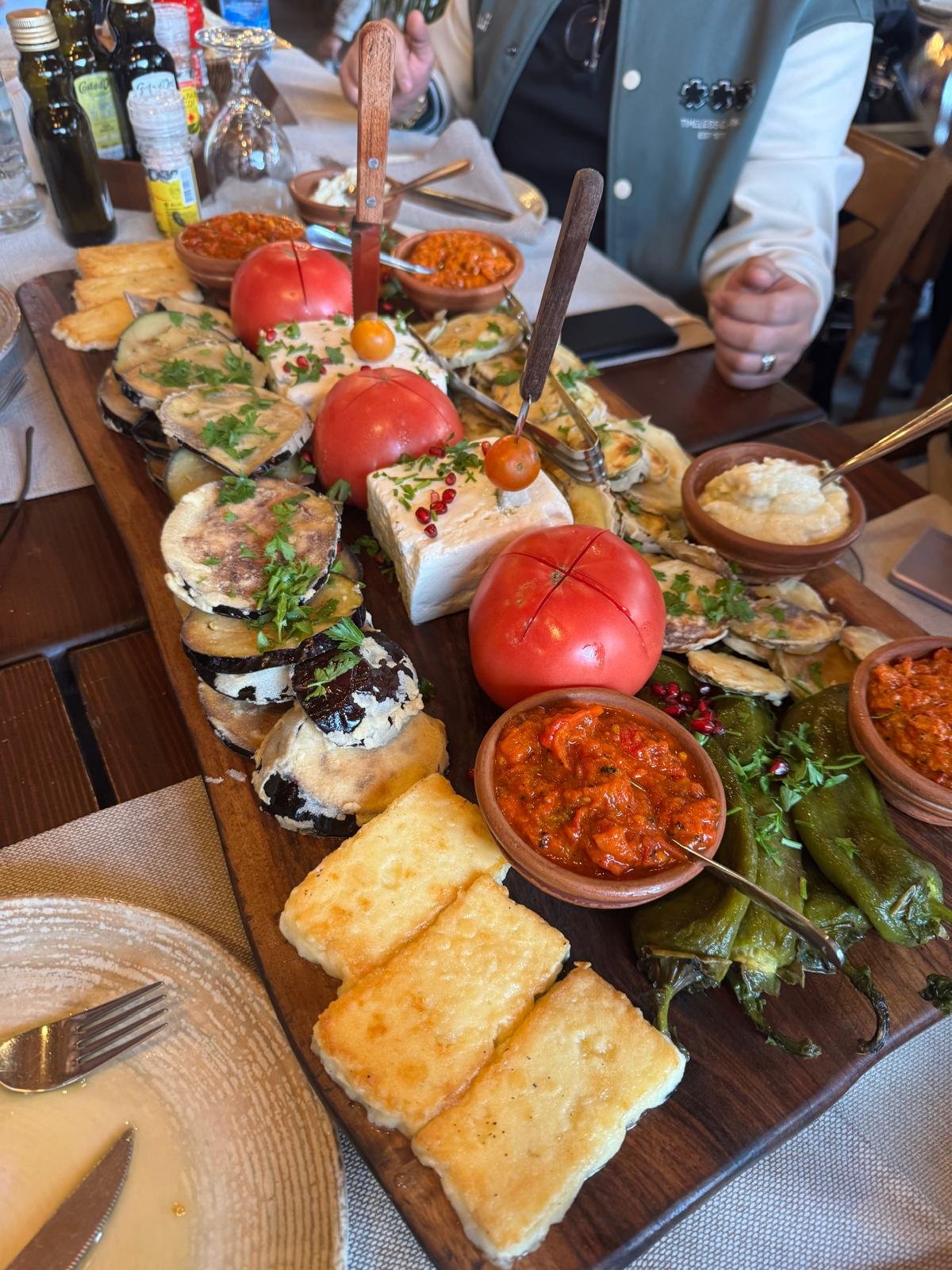 A wooden serving board with assorted Mexican appetizers, including sliced eggplant with herbs, baked cheese, stuffed jalapeños, fresh tomatoes, pickled vegetables, and small bowls of salsa, served on a dining table.