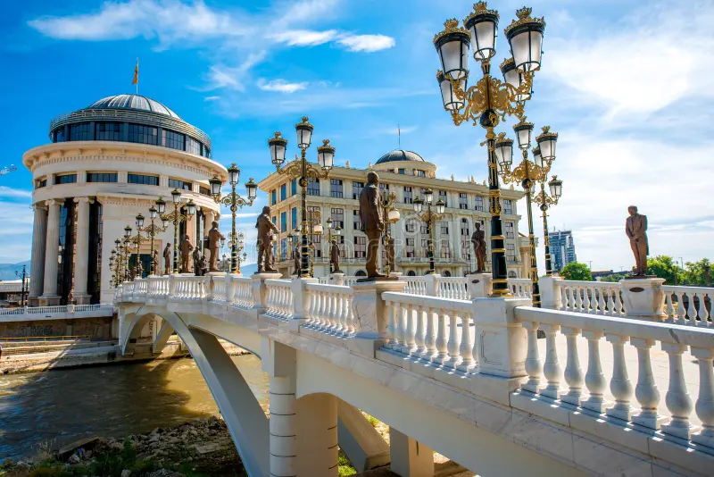 A decorative bridge with white railings and ornate street lamps leading to a classical-style building with a dome and sculptures, set against a blue sky with clouds.