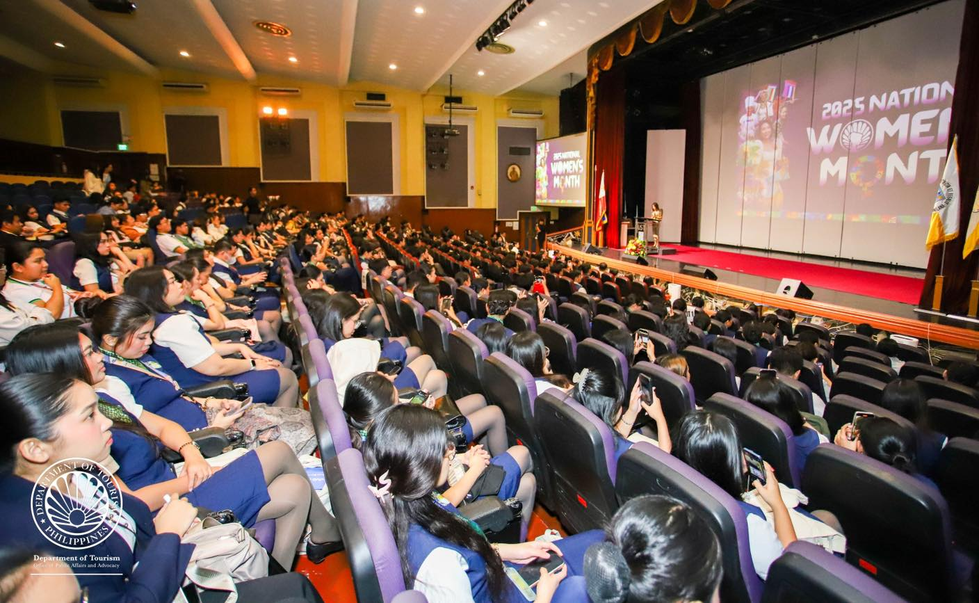 Large auditorium filled with students attending the 2023 National Women's Month event, with a stage and a speaker at the podium.