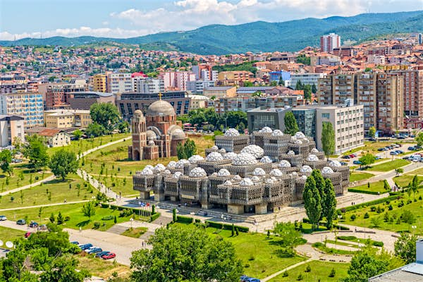 A cityscape with a historic Ottoman-style mosque surrounded by greenery and modern buildings against a backdrop of hilly terrain and blue sky.