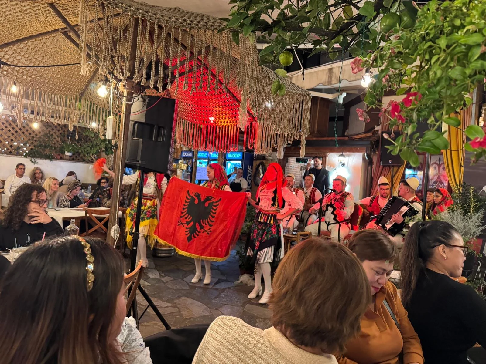 A lively indoor gathering with people enjoying traditional Albanian music and dance. Dancers in colorful, traditional Albanian folk costumes perform on a small stage, holding a red flag with a black eagle in the center. The audience is seated at tables, watching the performance amidst decorative lights, plants, and ethnic decor.