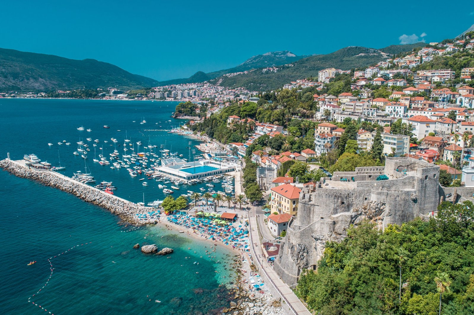A scenic coastal city with a harbor filled with sailboats and yachts, a beach with umbrellas, a historic stone fortress on a hill, and colorful buildings on lush green hillsides under a blue sky.