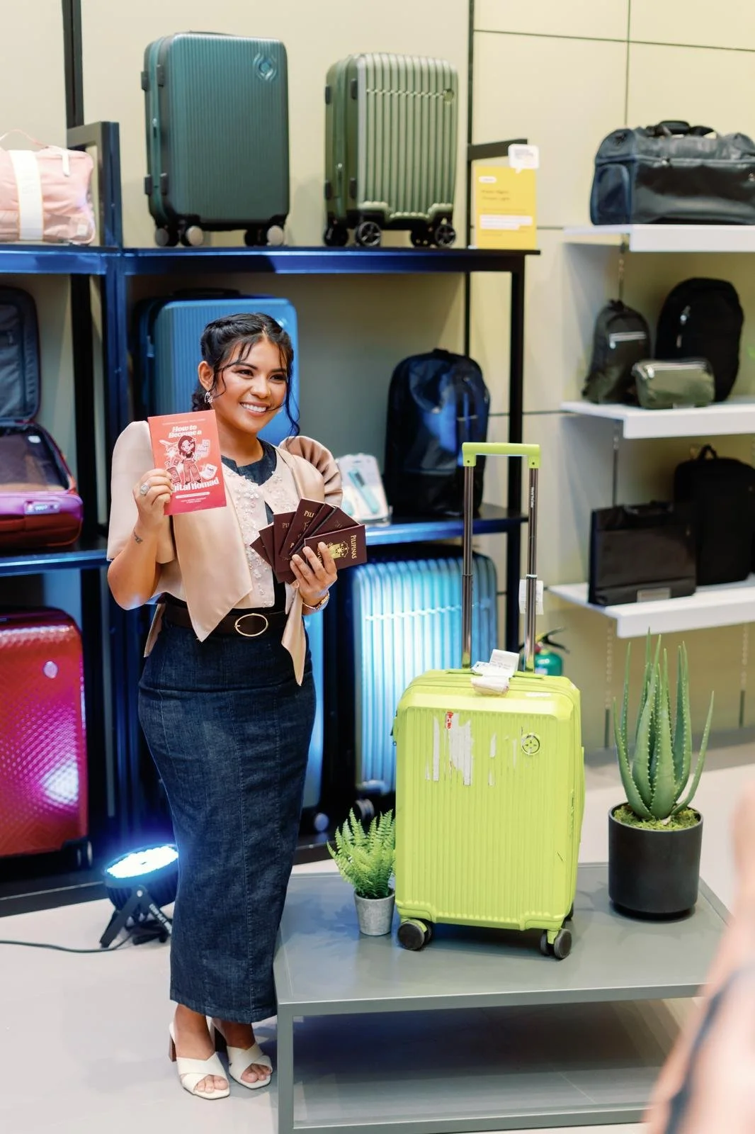 A woman smiling and holding travel brochures and passport in an airport luggage store, standing next to a bright yellow suitcase and potted plants.