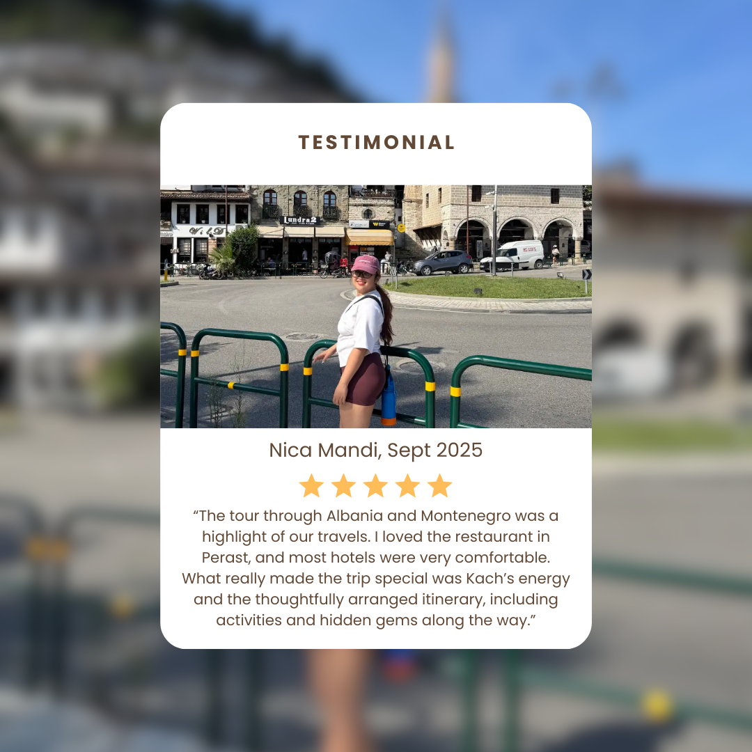 A woman in a pink cap and white shirt standing near green railings on a city street in Albania with buildings, cars, and pedestrians in the background.