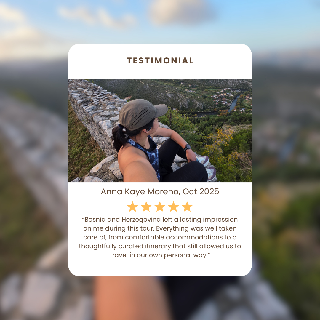A woman sitting on a stone wall overlooking a valley and river, wearing a cap and sporty clothing, taking a selfie.