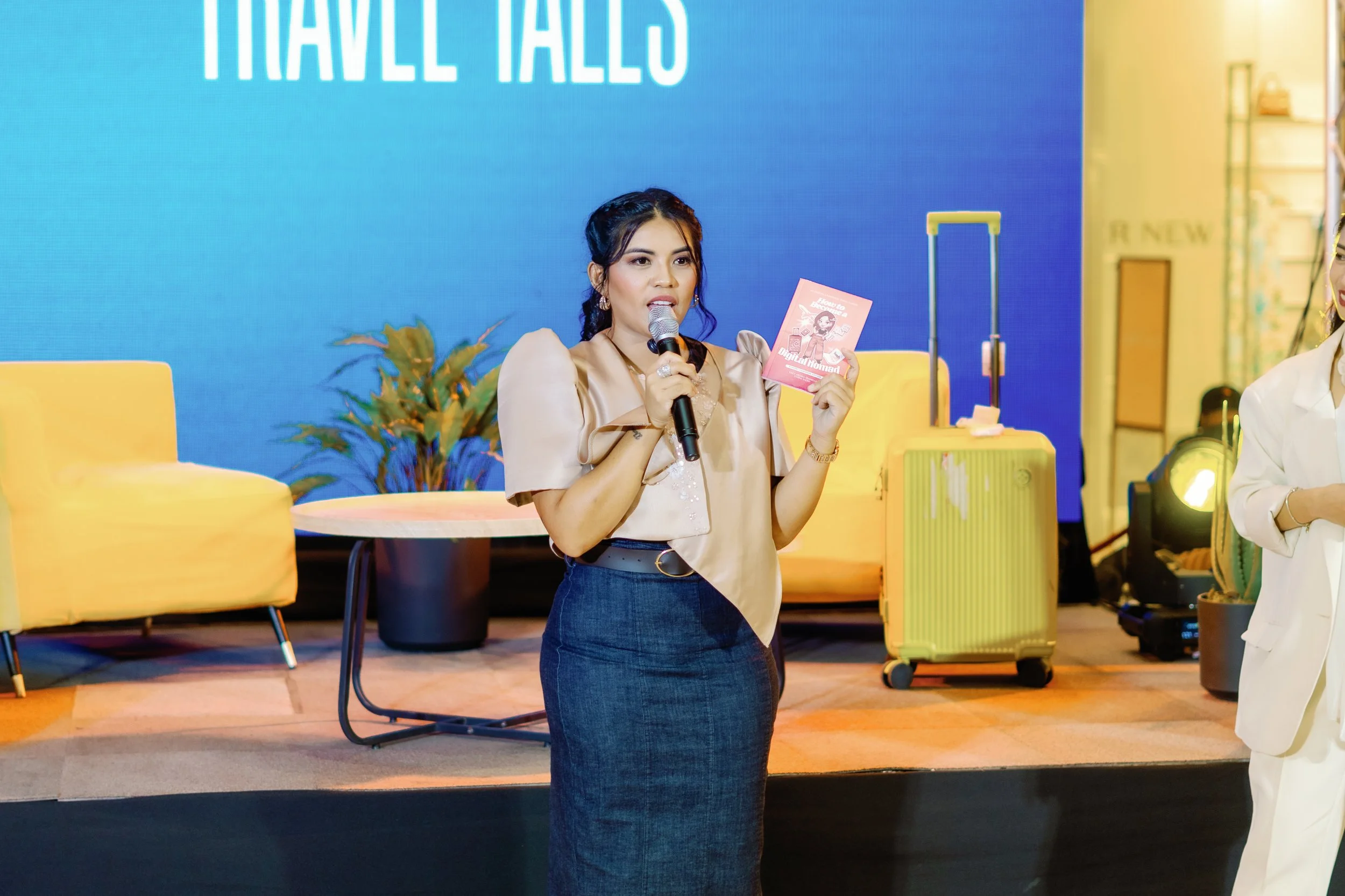 A woman speaking into a microphone while holding a pink booklet on a stage with yellow chairs, a table, and a yellow suitcase, with a large blue screen behind her that says "TRAVEL TALES."