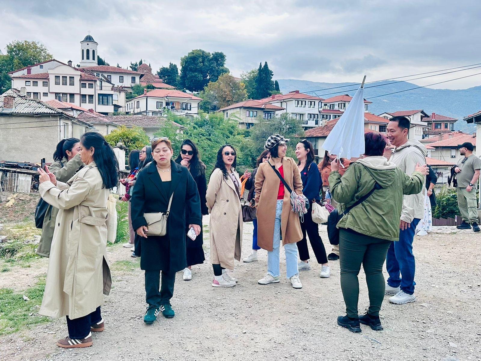 Group of people sightseeing and taking photos outdoors in a village with houses on a hillside and mountains in the background.