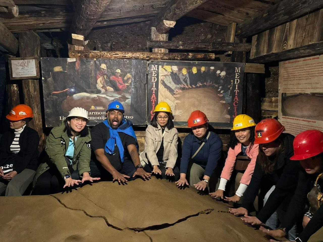 Group of people wearing safety helmets touching a large, cracked piece of rock in an indoor mining museum with informational posters on the wall.