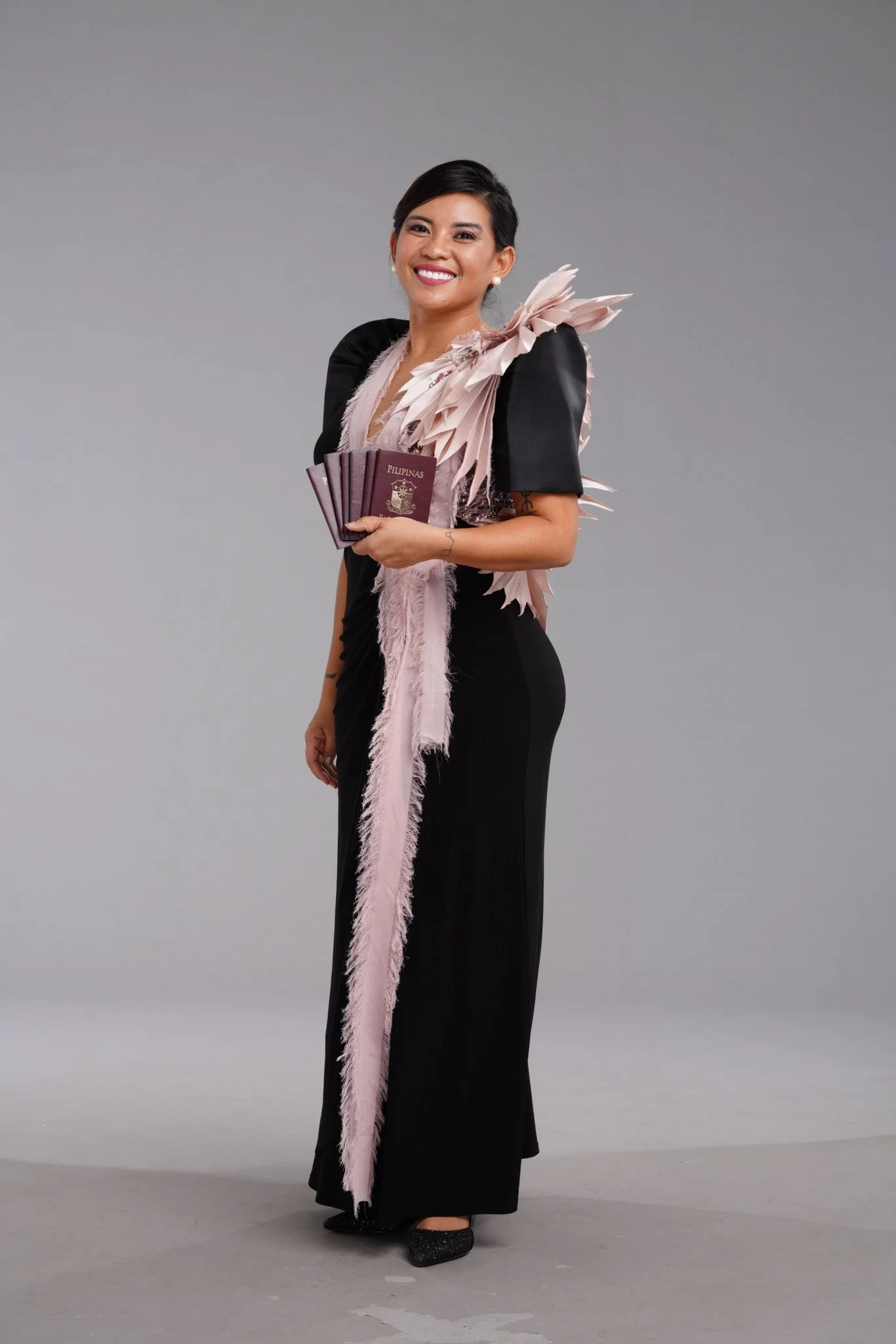 A woman in a black dress with decorative feathered accents on her shoulders and a matching feathered sash, holding several passports, standing against a plain background and smiling.