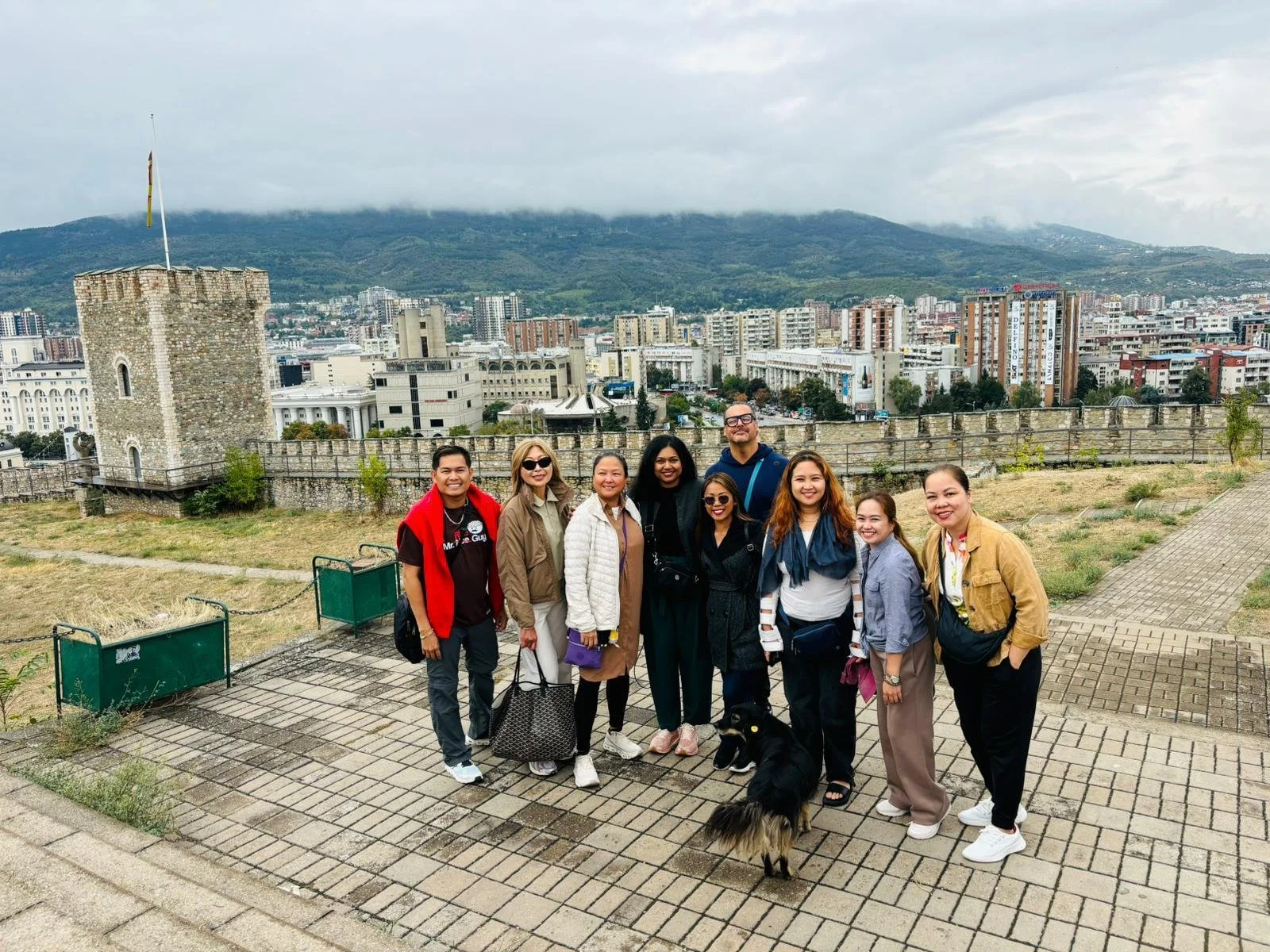 Group of ten people standing outdoors on paved area, smiling, with cityscape and mountains in background, and a black dog in front.