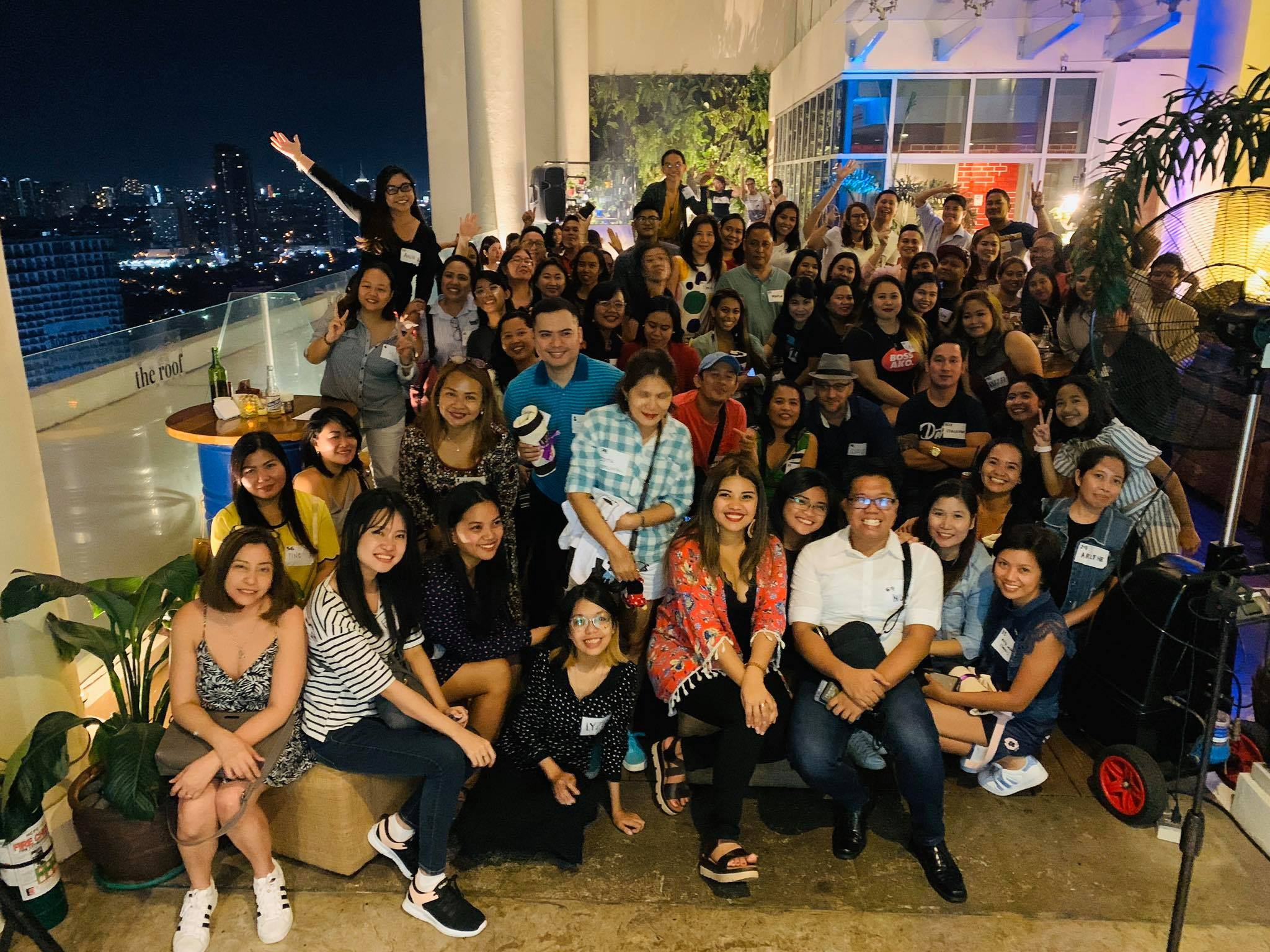A large group of people gathered on a rooftop at night, smiling and posing for a group photo. The city skyline with tall buildings and lights is visible in the background, and some people are holding drinks and making peace signs.