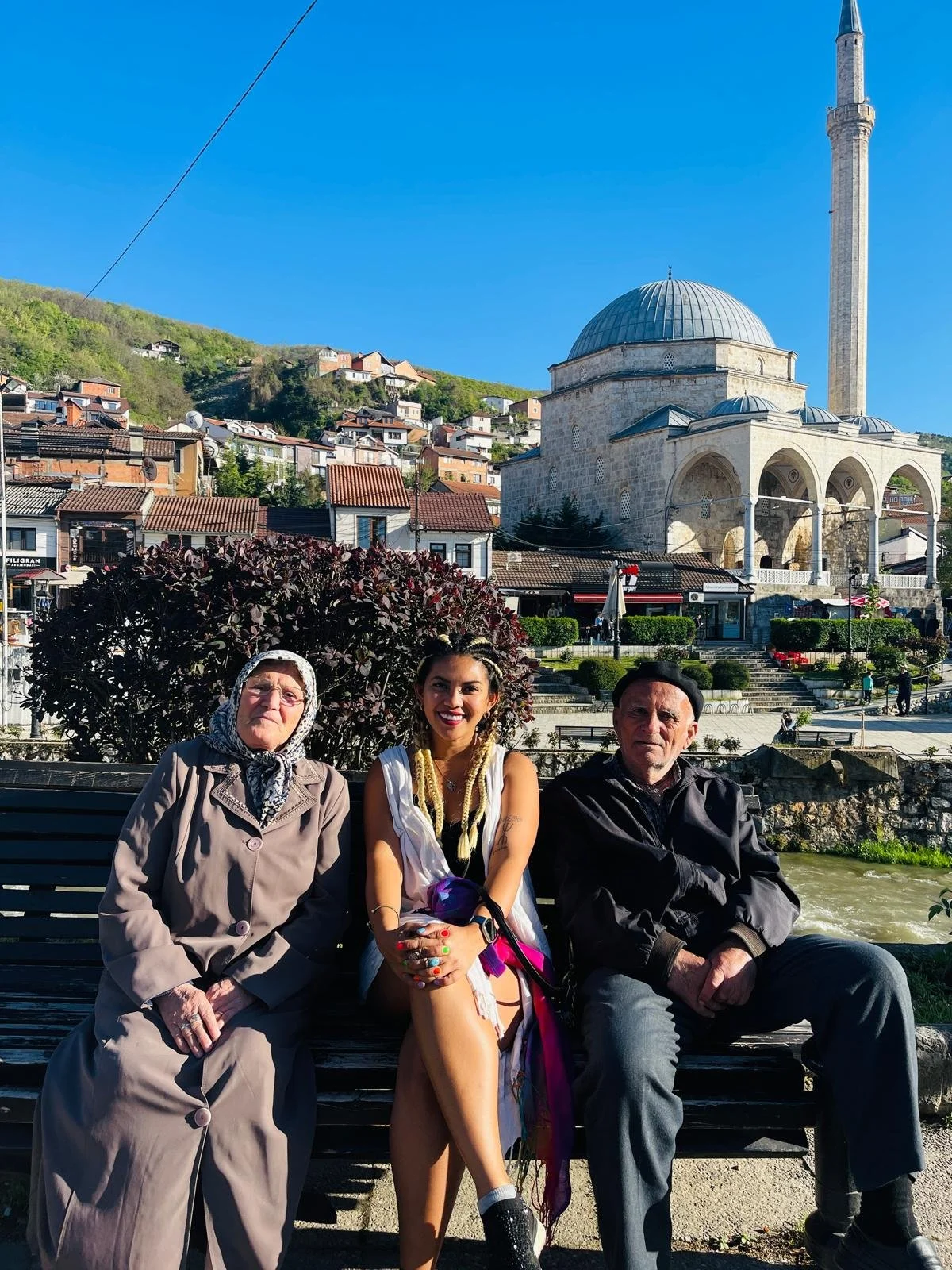 Three people sitting on a bench in front of a historic mosque with a large dome and minaret, hillside houses, and a clear blue sky.