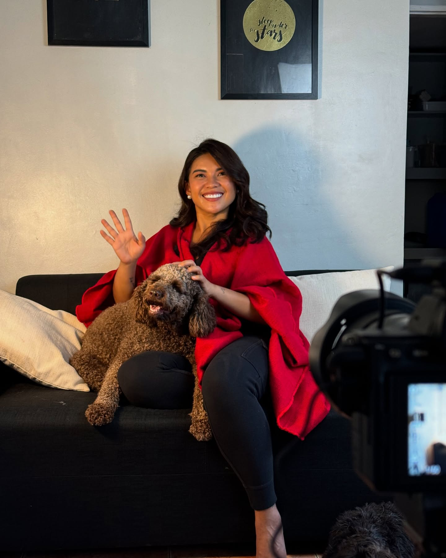 A woman in a red shawl sitting on a black couch with a brown poodle on her lap, smiling and waving at the camera in a living room with wall art.