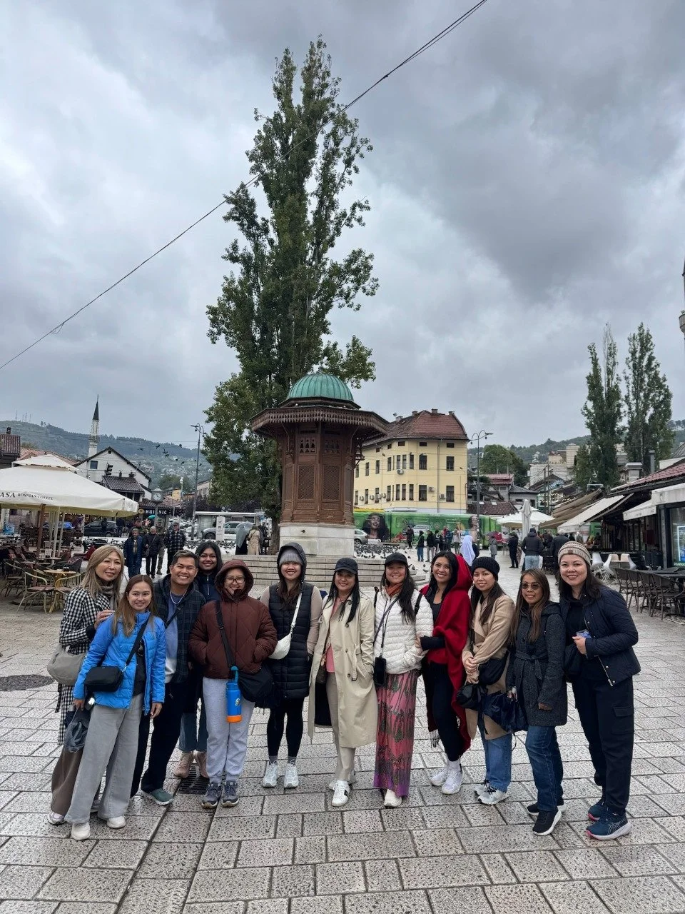 Group of twelve women and men standing on a paved square, smiling, in front of a historic metal kiosk with a green dome, surrounded by outdoor cafes, trees, and buildings, with a cloudy sky overhead.