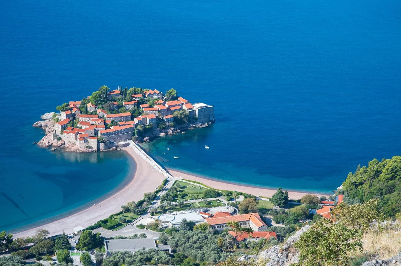 An aerial view of a small island with red-roofed buildings, surrounded by clear blue water, with a beach and green landscape in the foreground in Budva, Montenegro.
