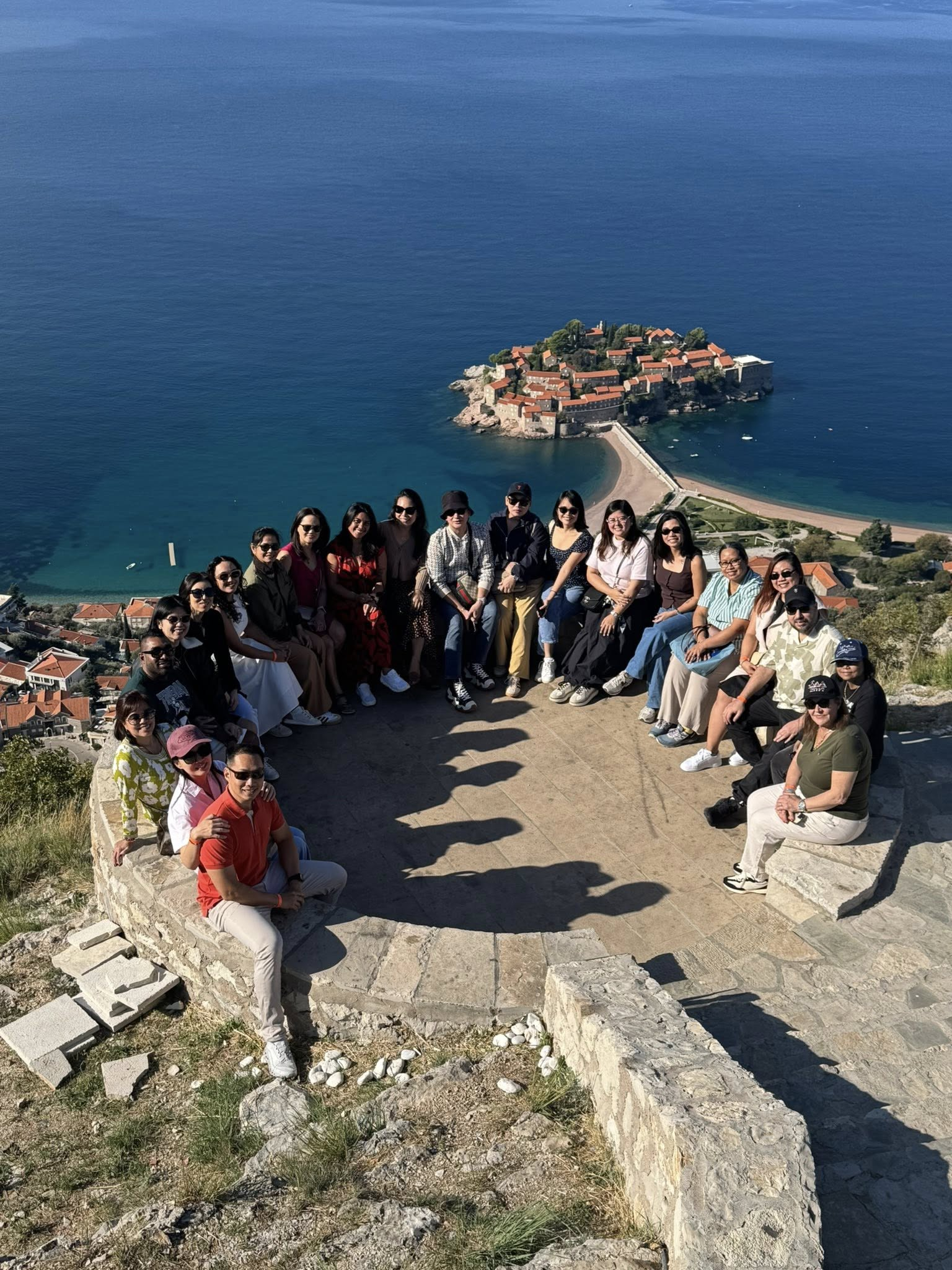 Group of people sitting on a stone ledge overlooking a coastal town and a small island in the sea.