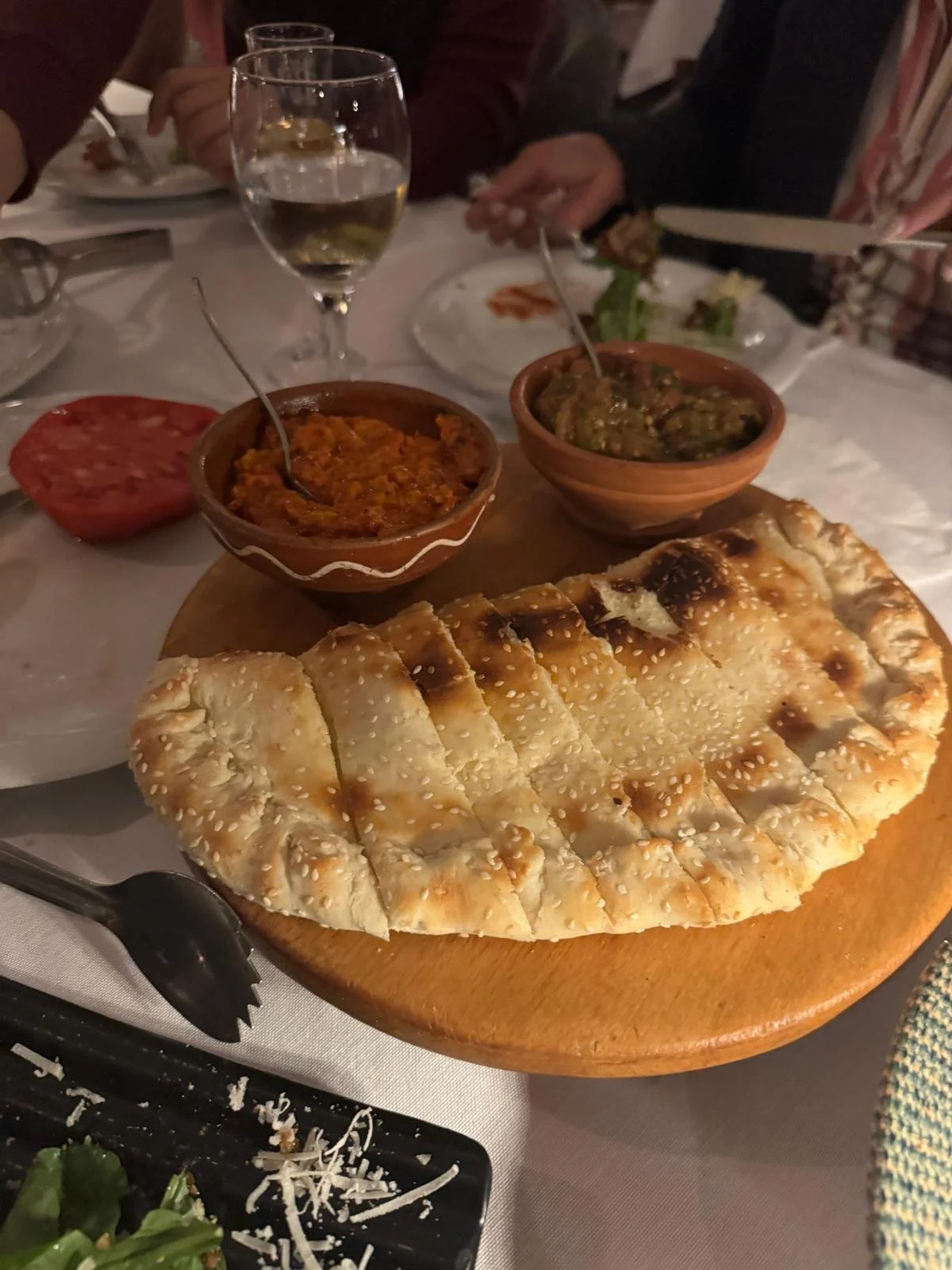 A large piece of naan bread, two small bowls of curry, a sliced tomato, a glass of white wine, and some salad on a dinner table.