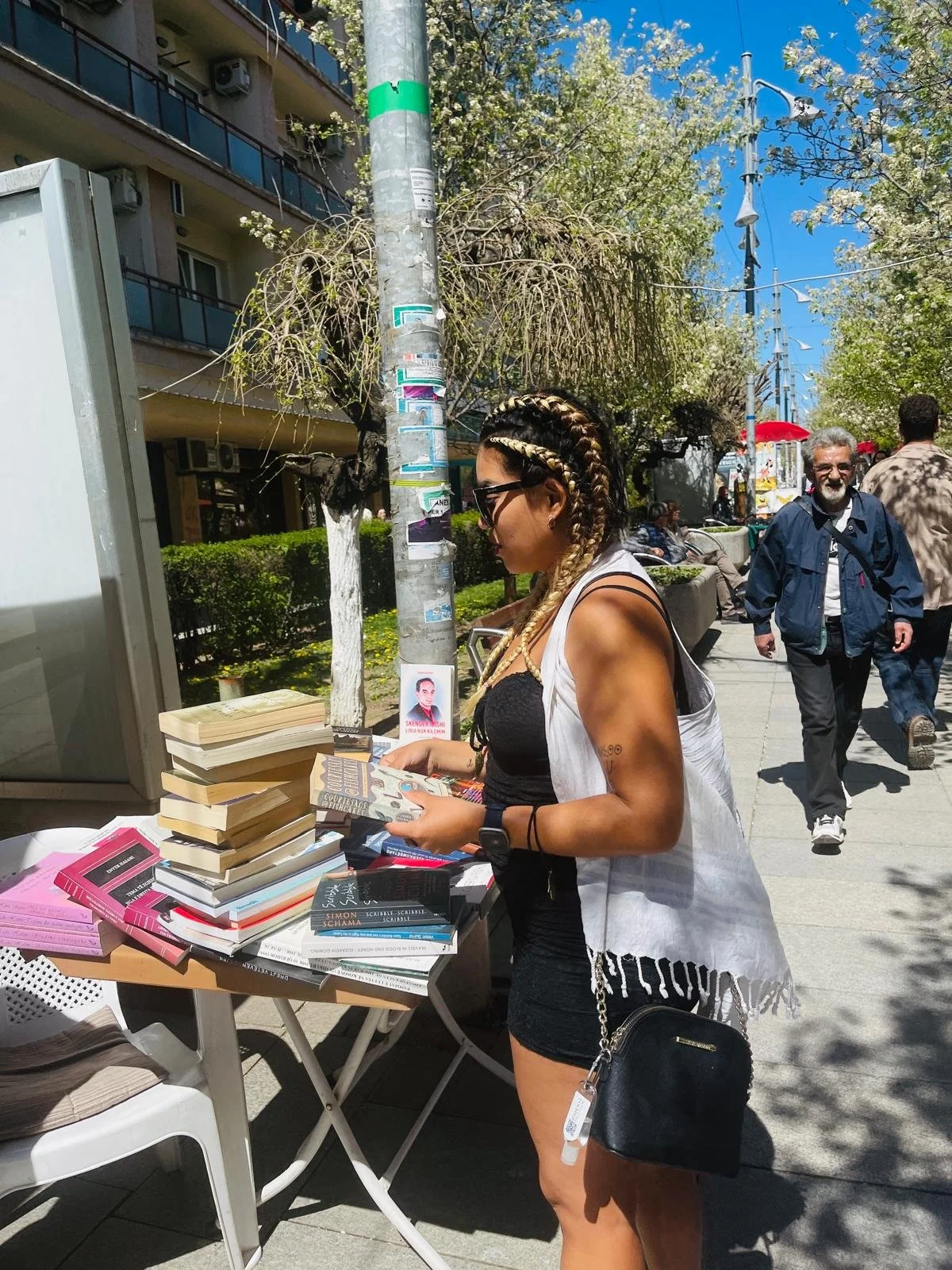 Woman browsing books at an outdoor book sale on a sunny day, with a table of books and pedestrians walking by in the background.