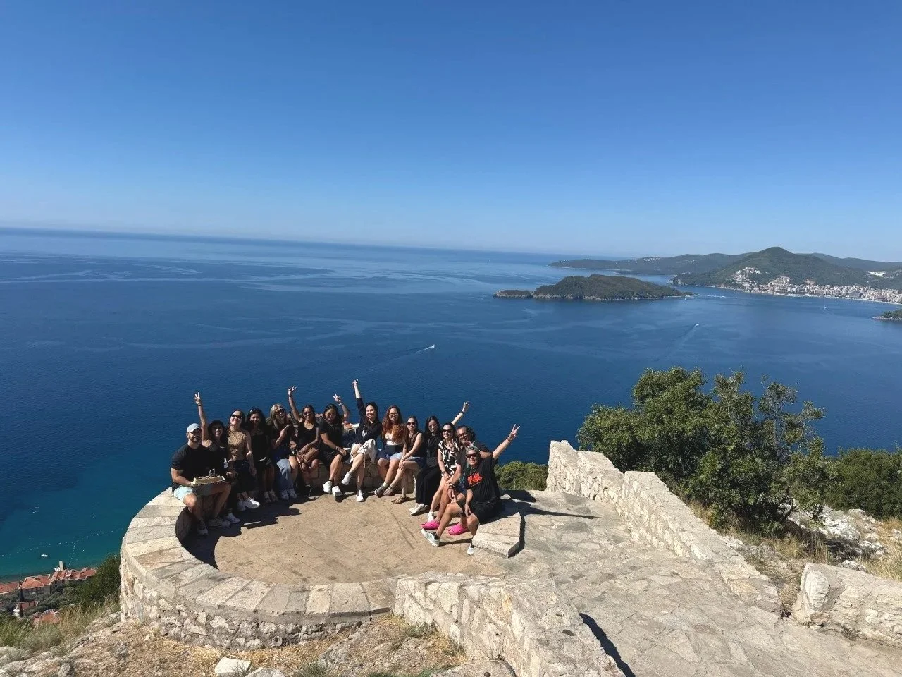 Group of people sitting on a stone platform on a hillside overlooking an expansive blue sea with islands and a coastal town in the distance, under a clear blue sky in Budva, Montenegro.